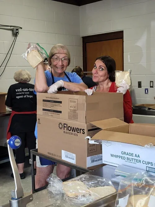 Two women smiling at a kitchen counter, packing bread and whipped butter cups into boxes, with several boxes of baking supplies around them.