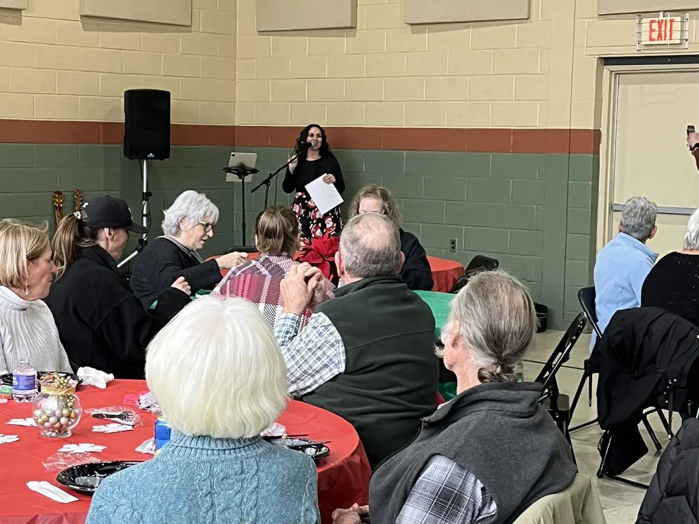 A woman speaking at a microphone during a gathering or event, with seated attendees at decorated tables, in an indoor hall with beige and green walls.