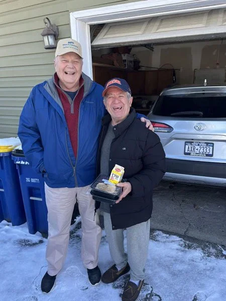 Two smiling men standing outside a garage, one holding a container of food and the other with an arm around him, with trash cans nearby and snow on the ground.