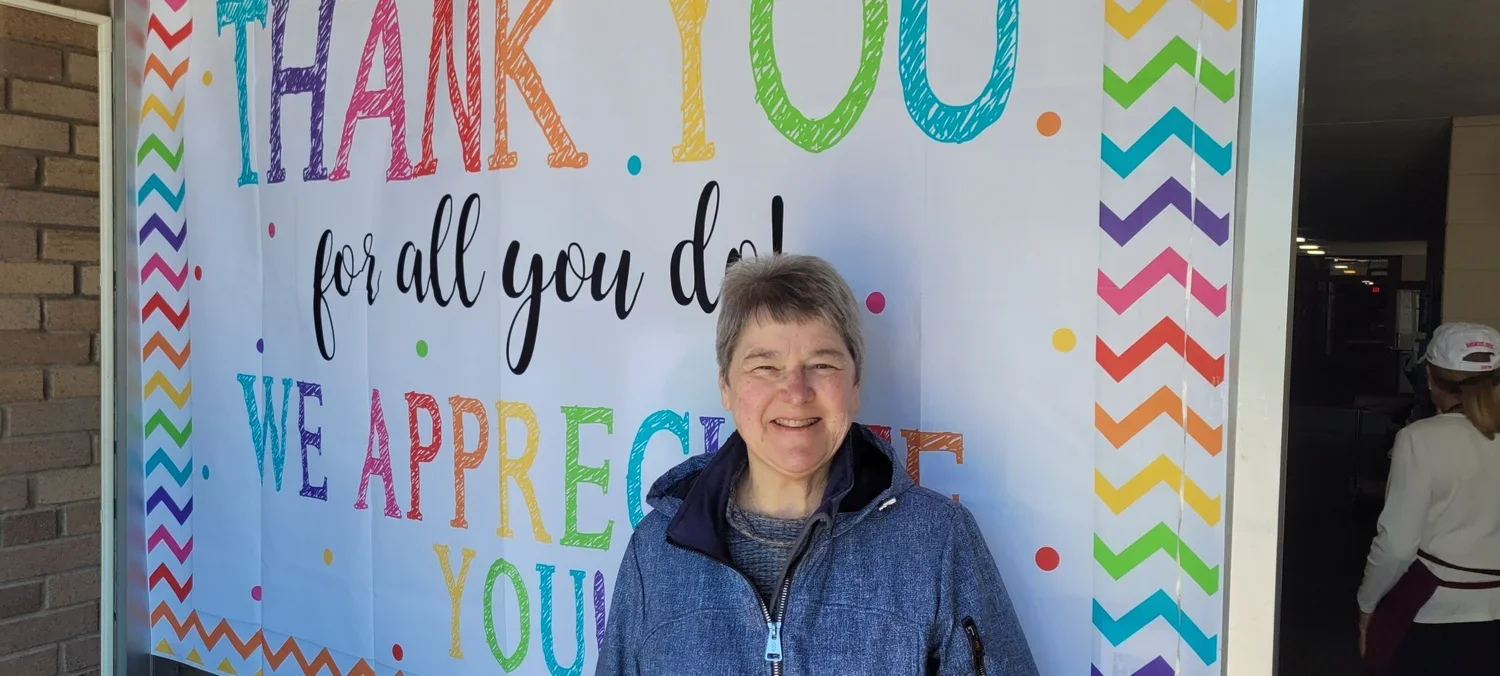 A smiling woman standing in front of a colorful thank you sign with rainbow and zigzag border.