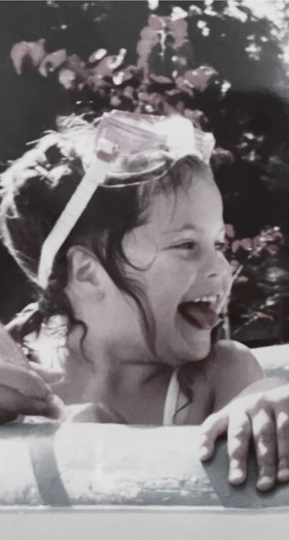 A young girl with wet hair and a big smile on her face, leaning on a swimming pool's edge, with a small plastic bucket on her head.