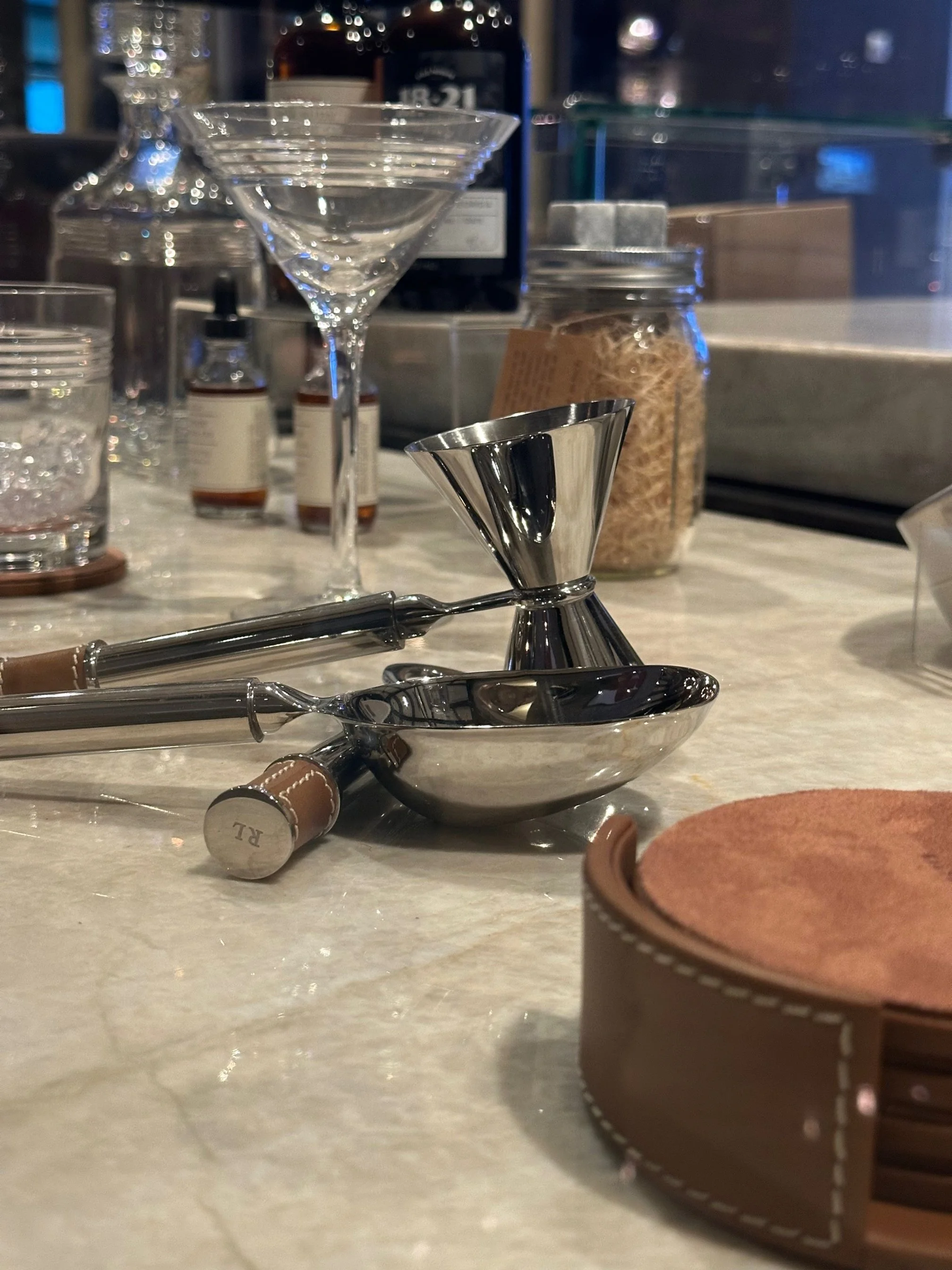Bar countertop with cocktail tools including a strainer, a jigger, and a spoon, along with various bottles and jars in the background.
