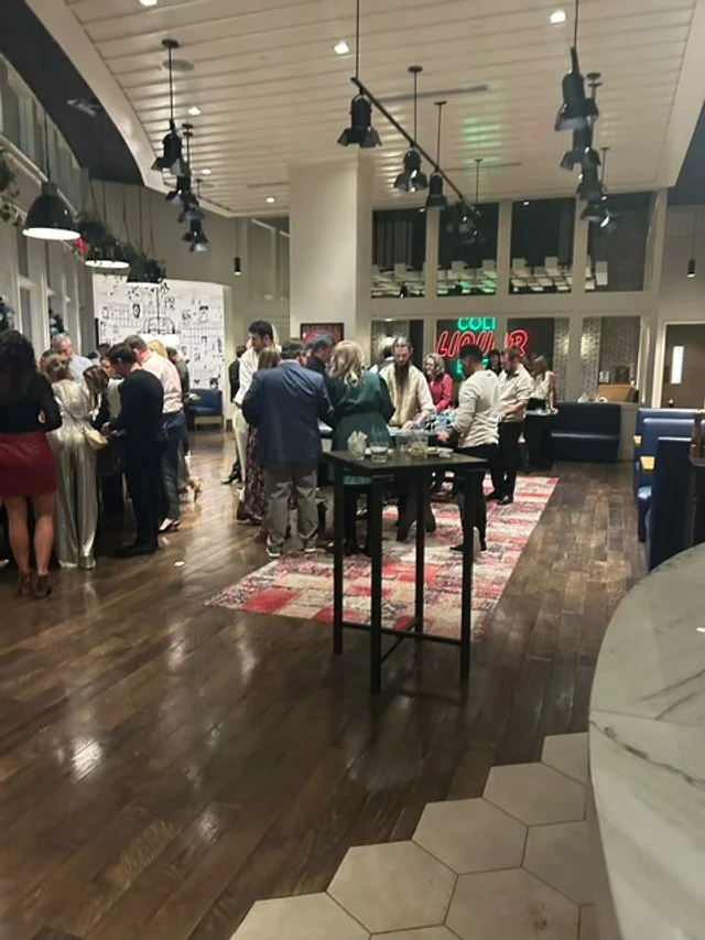 People gathered around a bar or service counter at a social event in a modern, well-lit indoor space with wooden floors and contemporary decor.