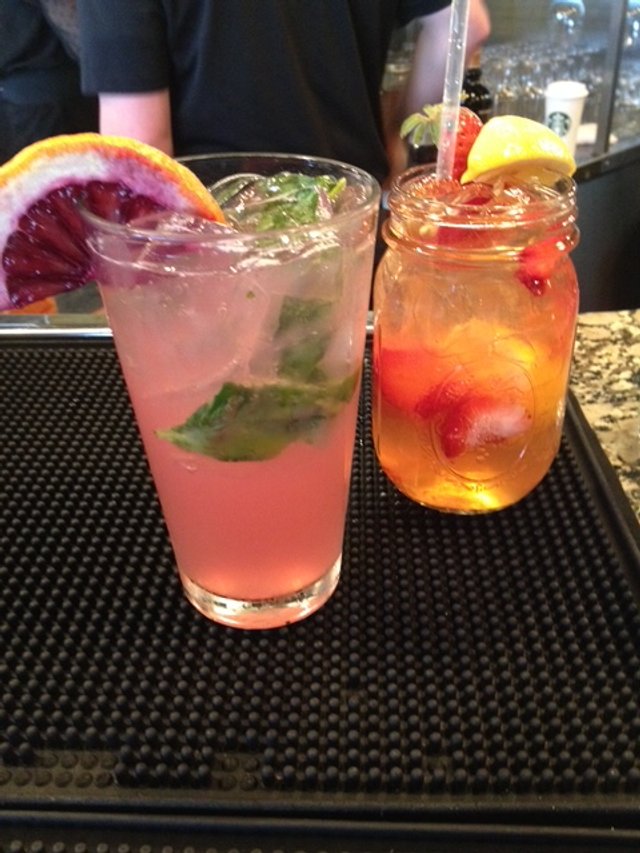 Two colorful cocktails on a black rubber bar mat. The left drink is pink with a slice of blood orange and mint leaves, and the right drink is orange with berries and a lemon wedge.