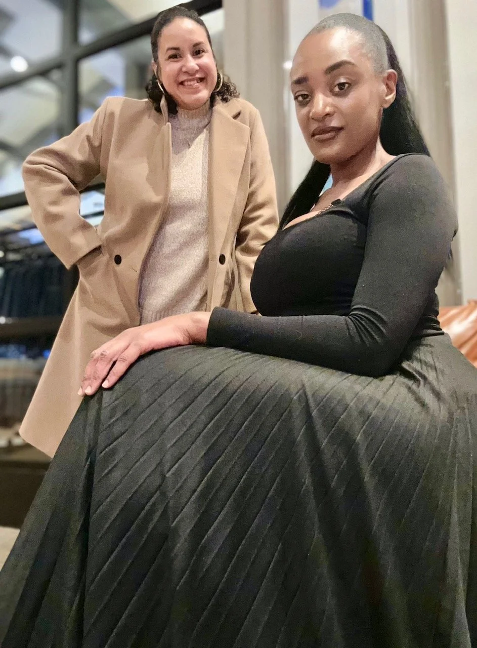 Two women posing indoors, one standing and smiling, the other seated with her arm resting on her lap, both dressed stylishly.