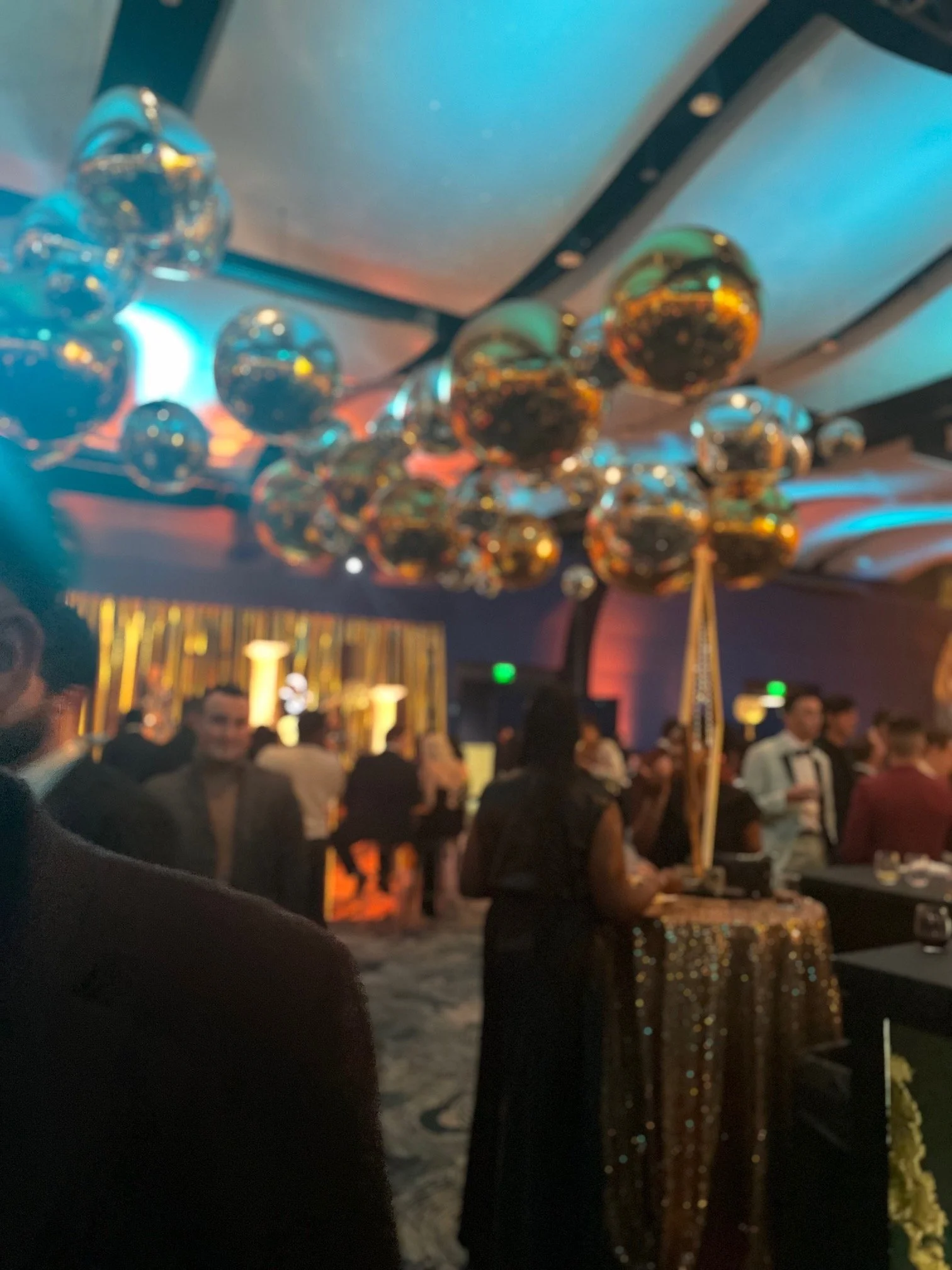 A party or celebration with gold and silver balloon decorations on the ceiling, a glittering gold tablecloth, and a crowd of people in formal attire in a dimly lit room with colorful lighting.