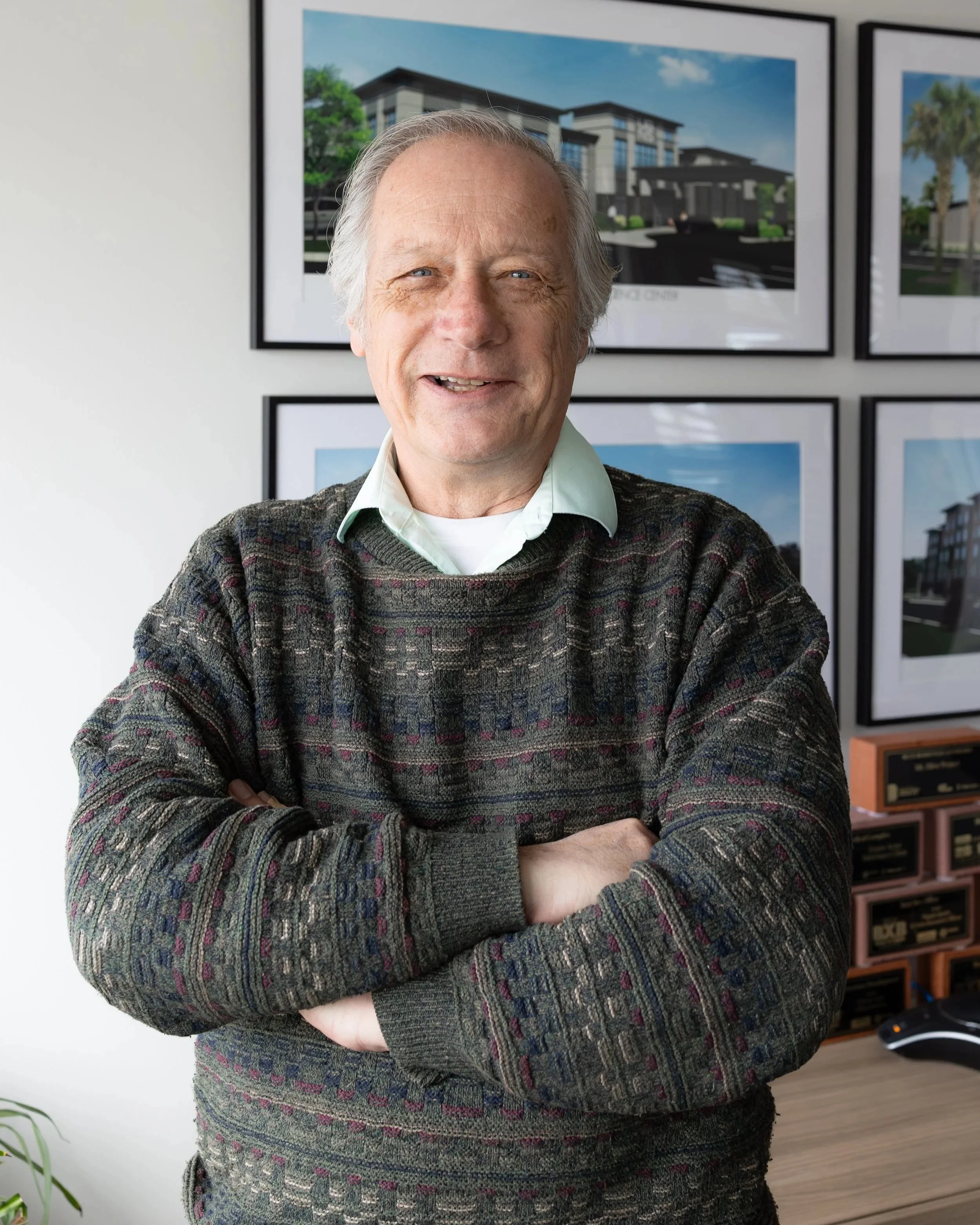 A man with dark curly hair and a beard smiling while leaning against a metallic wall, wearing a navy blue jacket, gray sweater, white shirt, and a smartwatch.