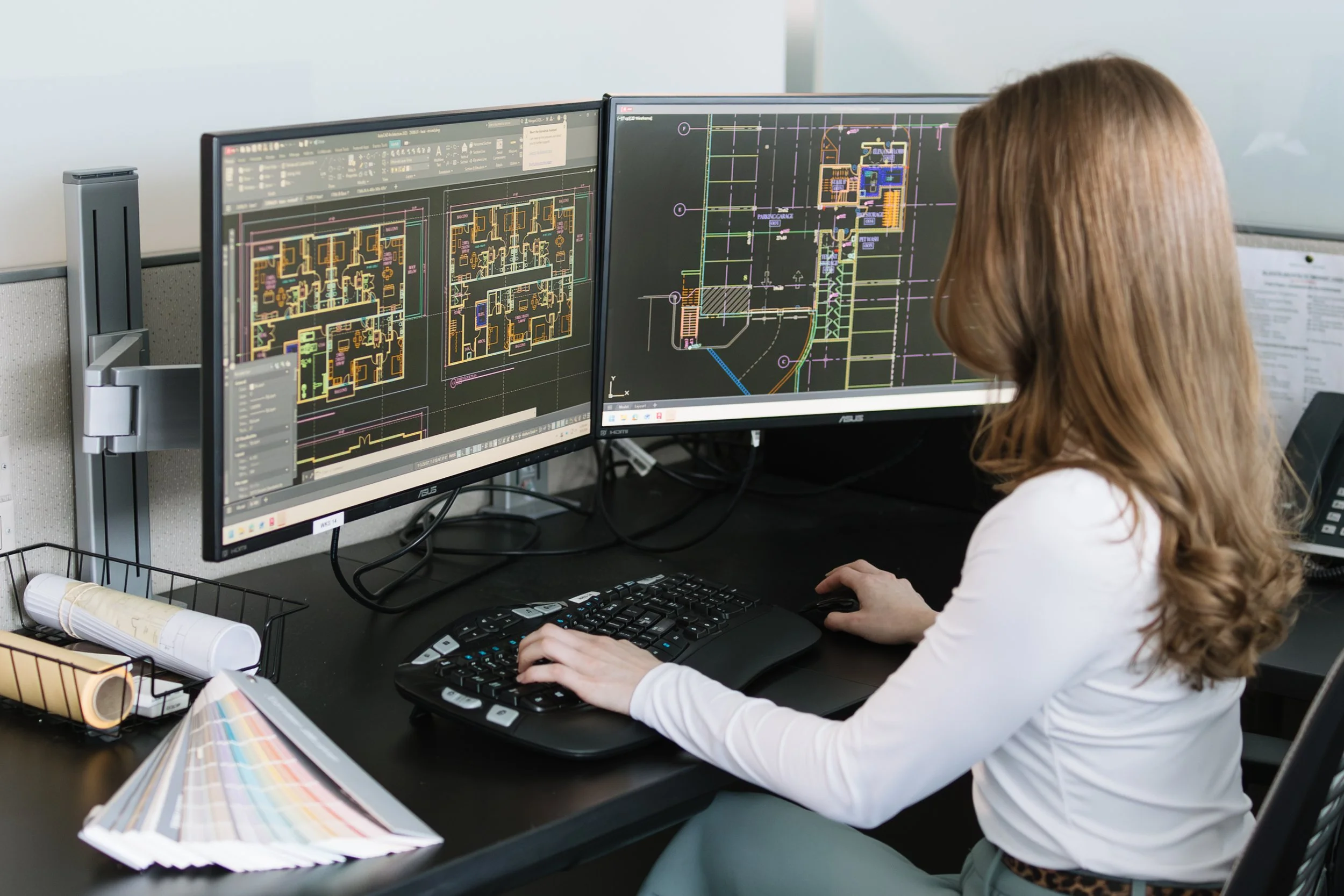 Woman working on architectural or engineering plans on dual computer monitors, with drafting tools and color samples on her desk.