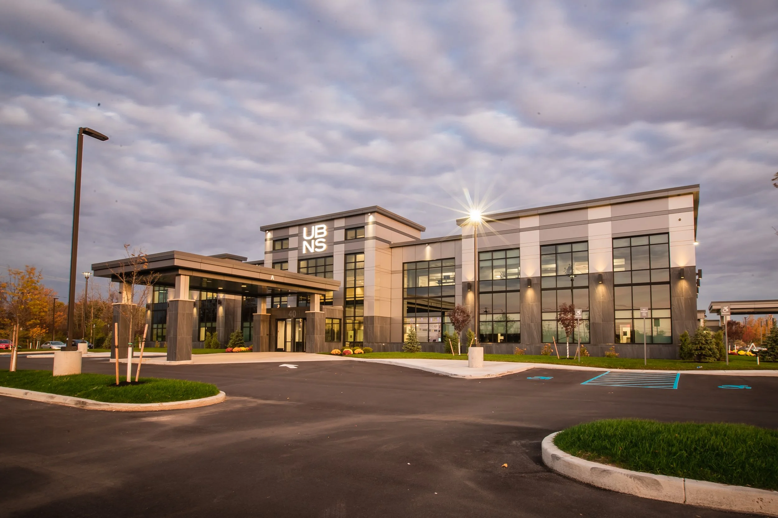 Modern hospital building with large glass windows and illuminated signage that reads "UBNS." Parking lot in foreground with marked accessible parking spaces and landscaped areas, under a partly cloudy sky.