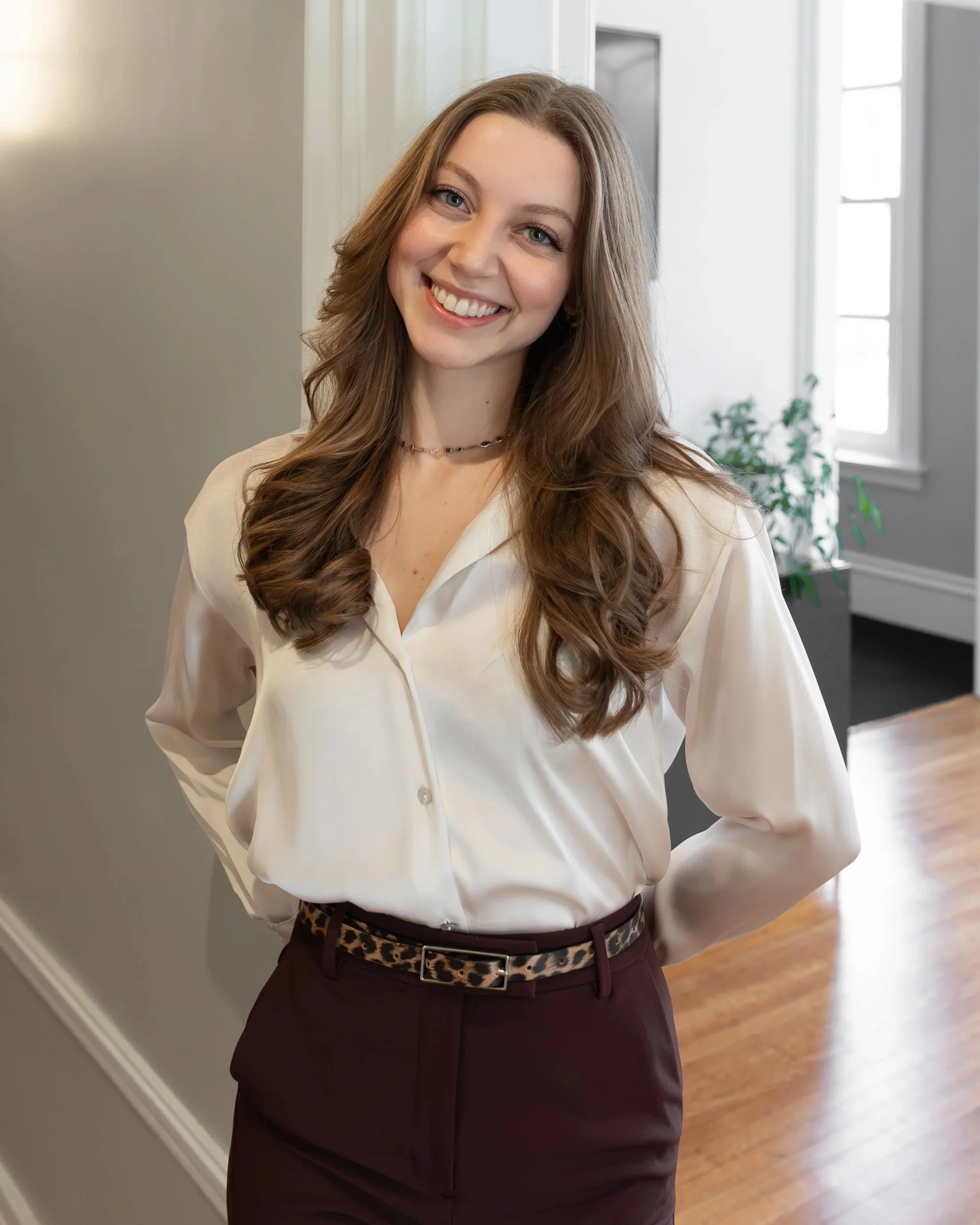 A young woman with long wavy brown hair and blue eyes smiling, wearing a cream satin blouse and maroon pants with a leopard print belt, standing indoors near a doorway with a window in the background.