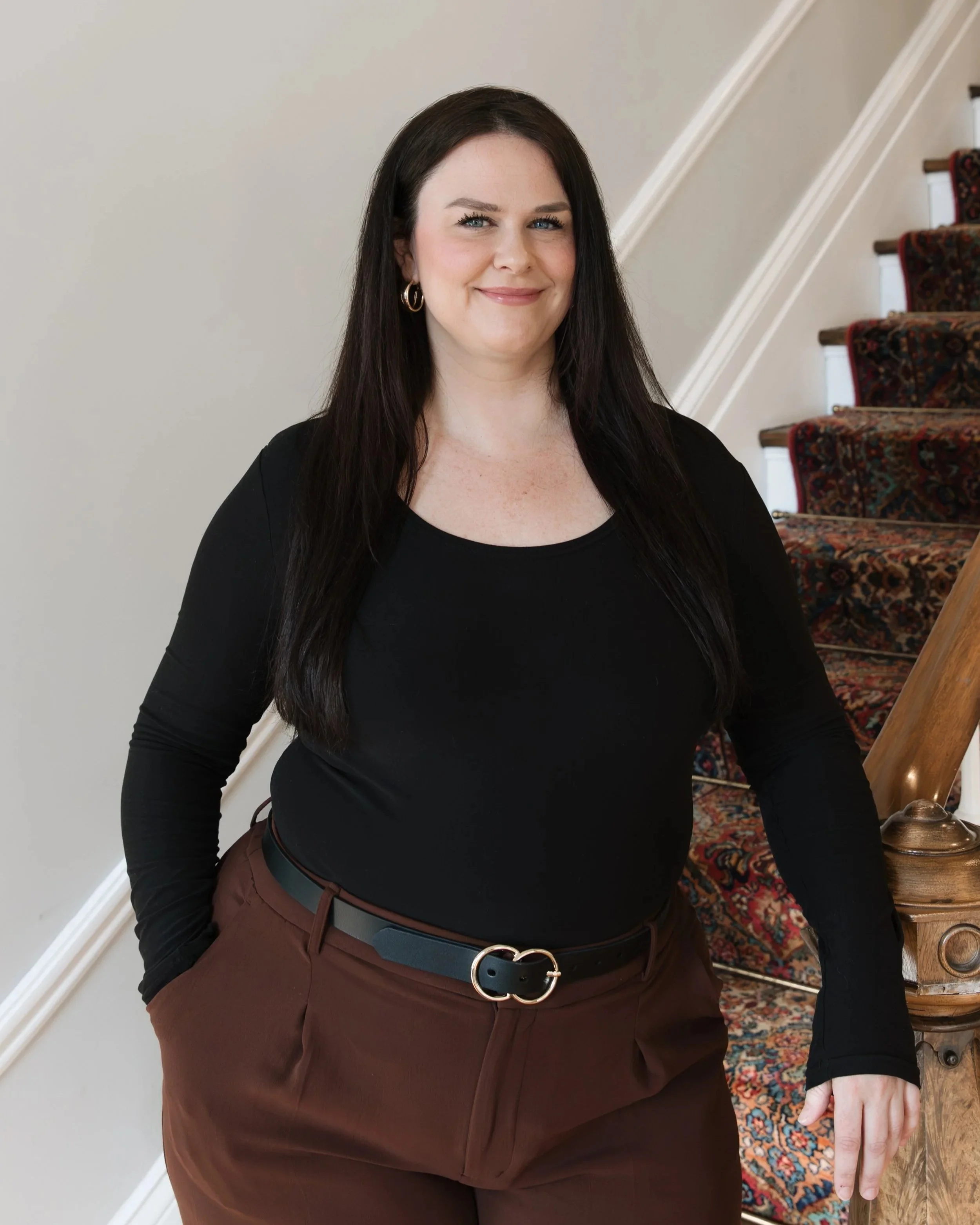 A woman with long dark hair, wearing a black top and brown pants, standing by a staircase with a carpeted runner, indoors.