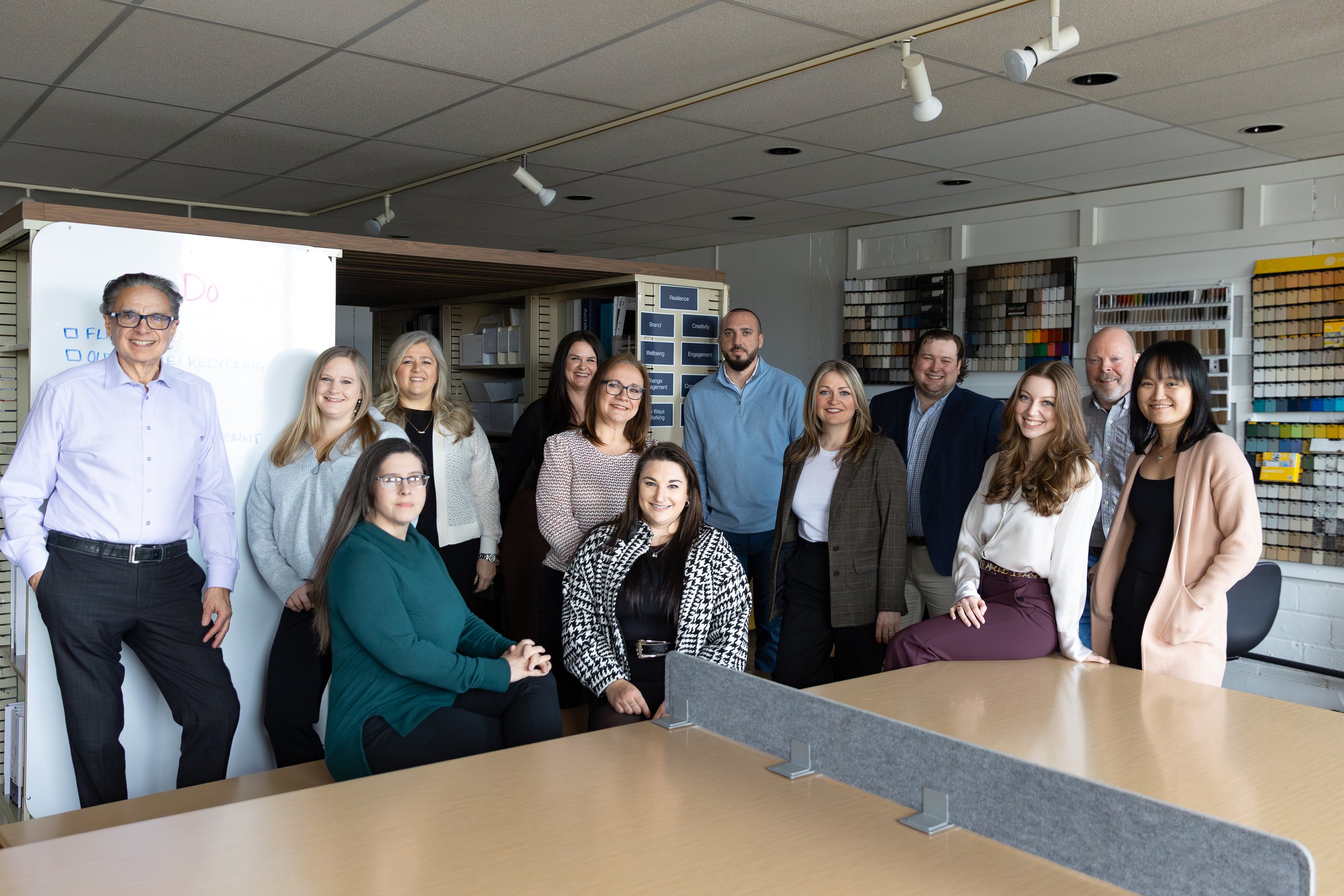 Group of 14 professionals in an office, some standing and some sitting, smiling at the camera with office supplies and shelves in the background.