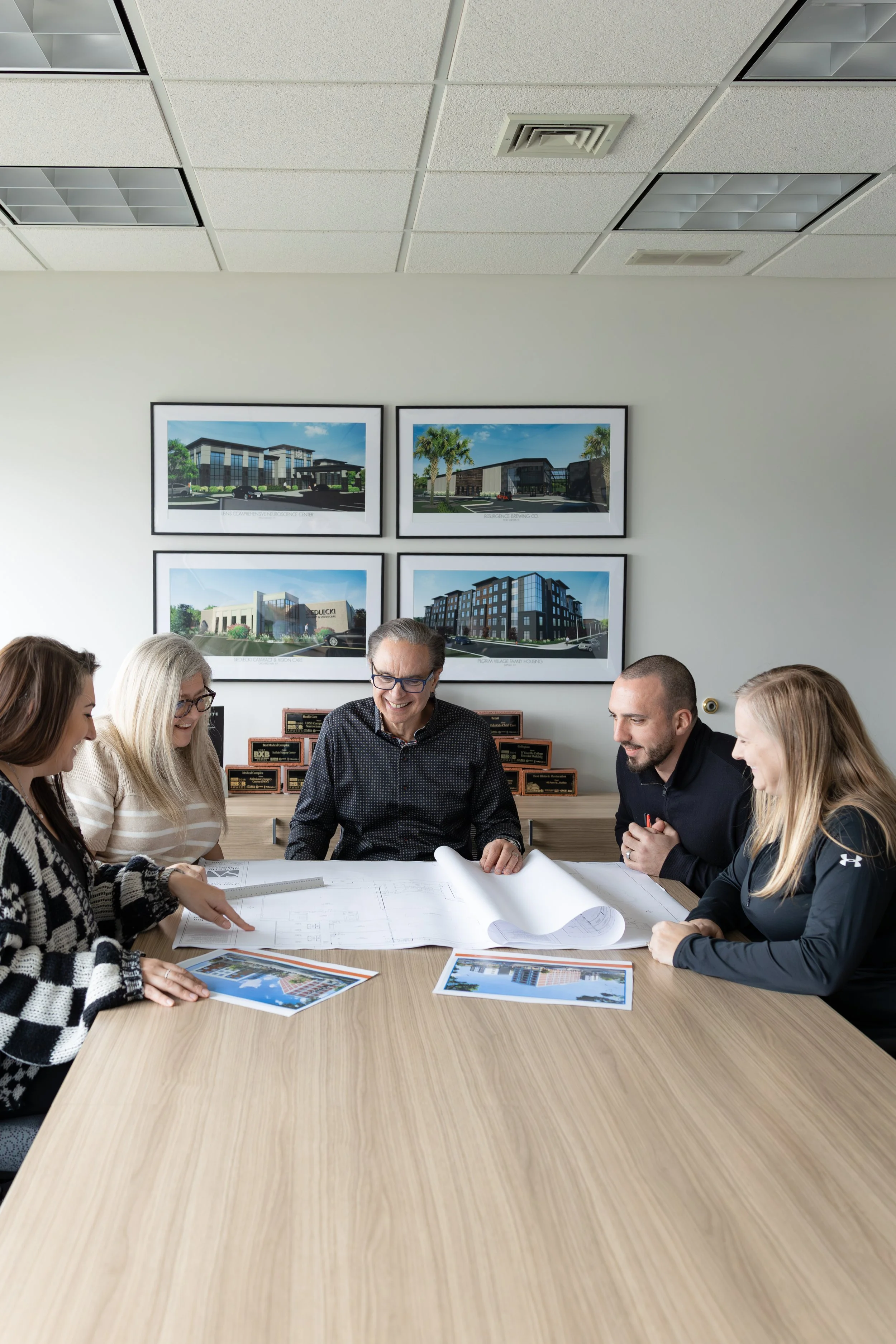 Group of five people sitting around a table looking at architectural blueprints and photos, in a conference room with framed building designs on the wall.