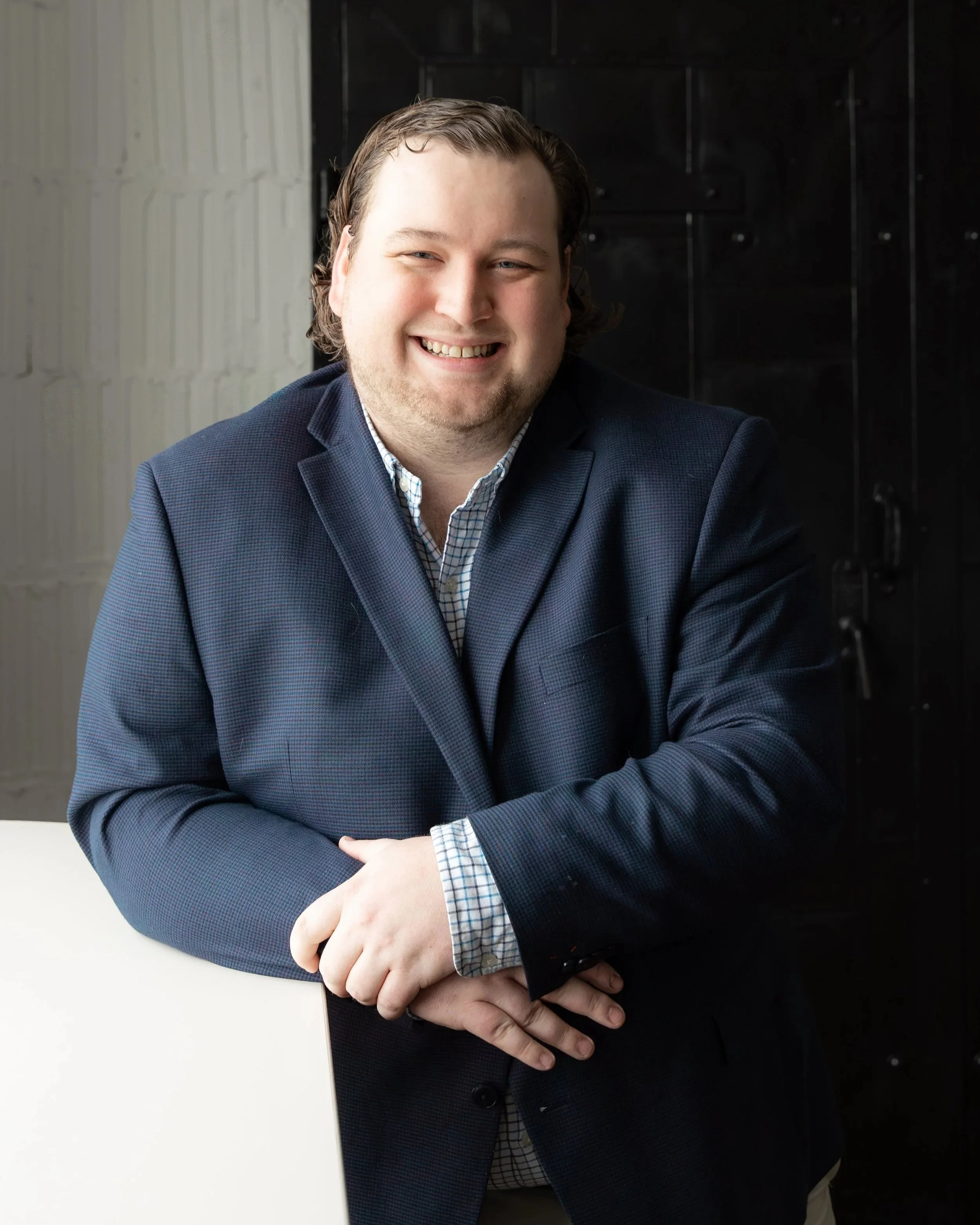 A man with light skin and brown, slightly curly hair, smiling, wearing a dark blue blazer and a checkered shirt, leaning on a white surface in front of a dark and light background.