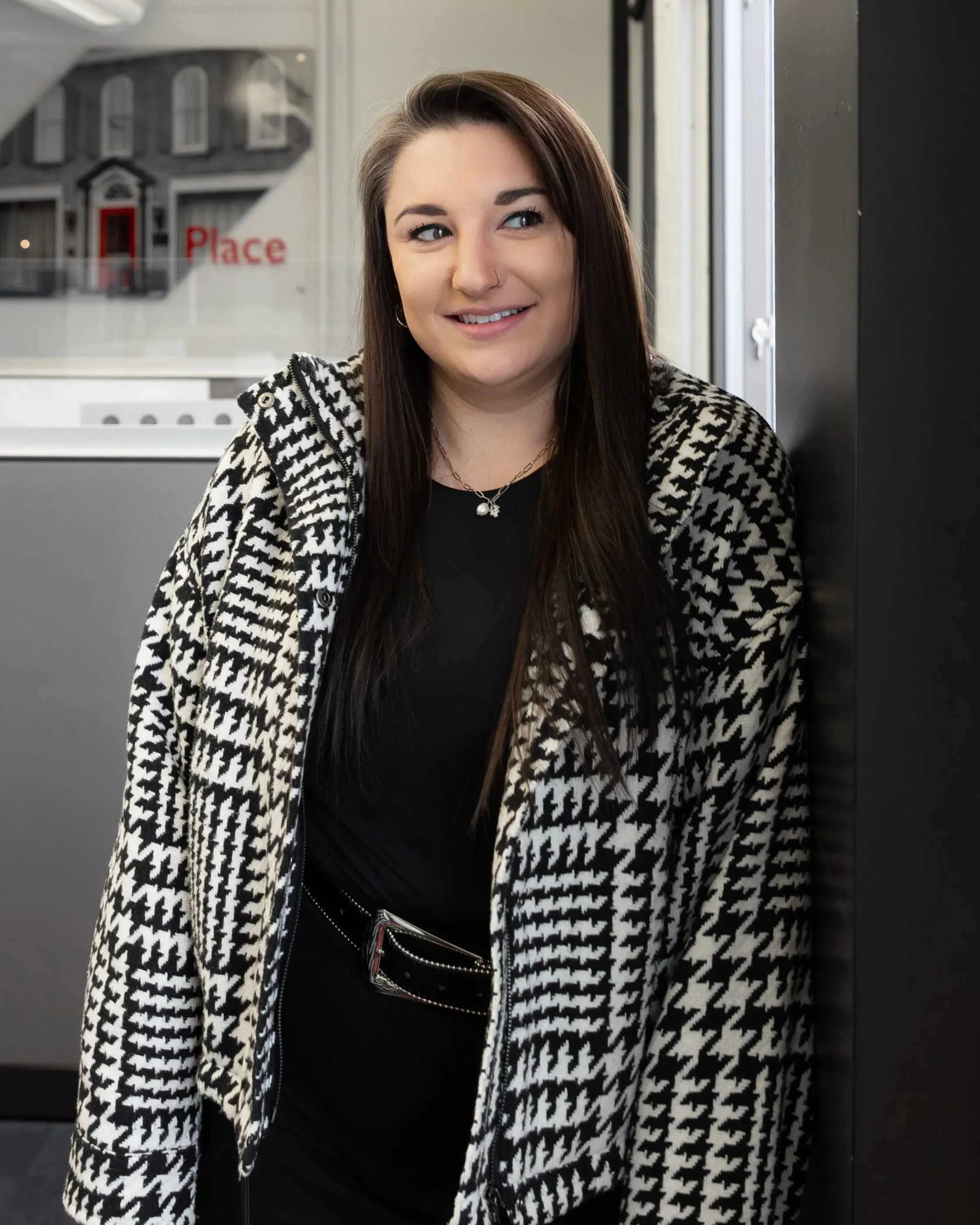 A woman wearing glasses and hoop earrings sits at a desk writing with a red pen, with a smartphone on the desk and a computer monitor in the background.