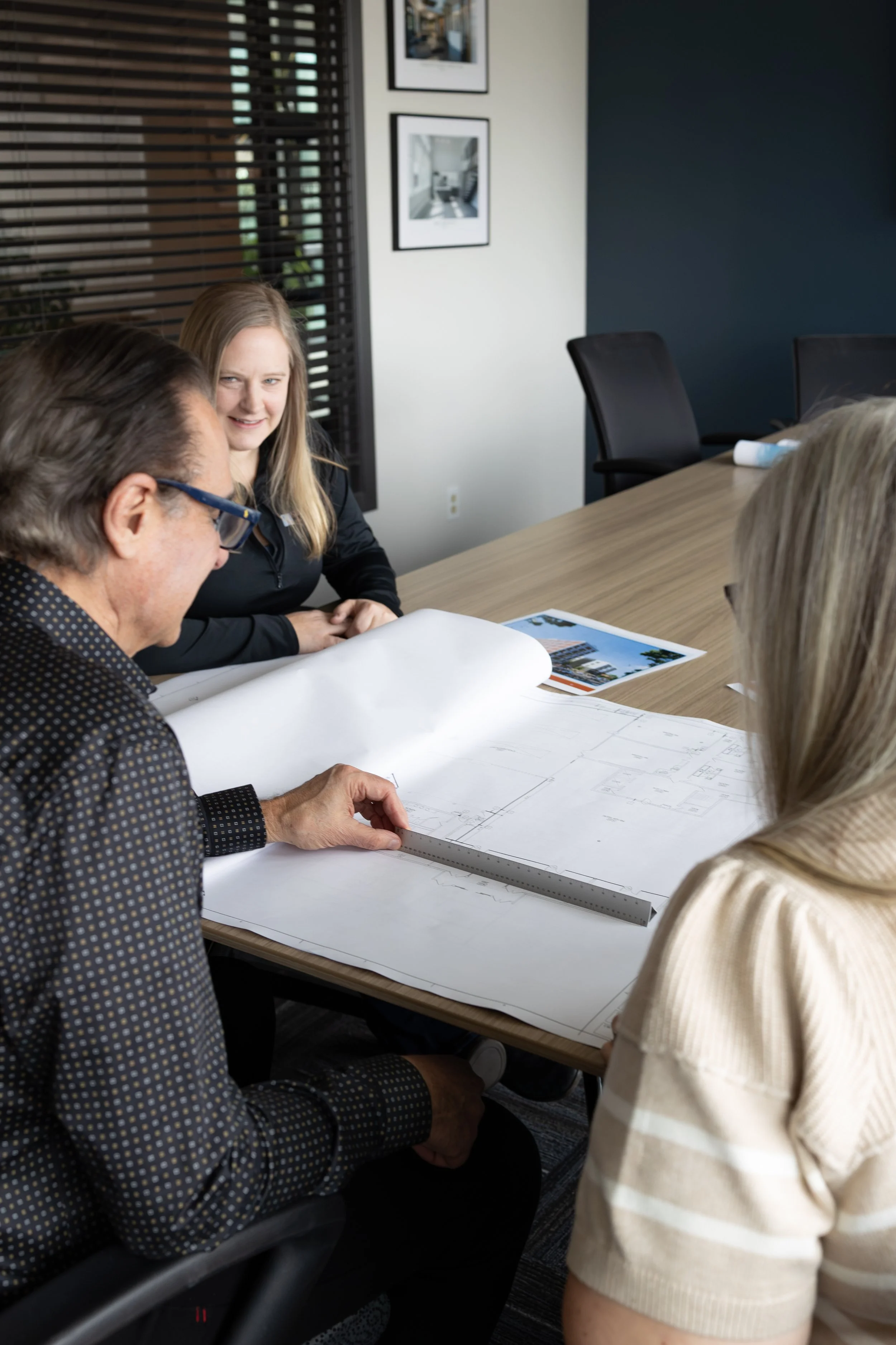 Three people are gathered around a conference table reviewing large architectural blueprints.
