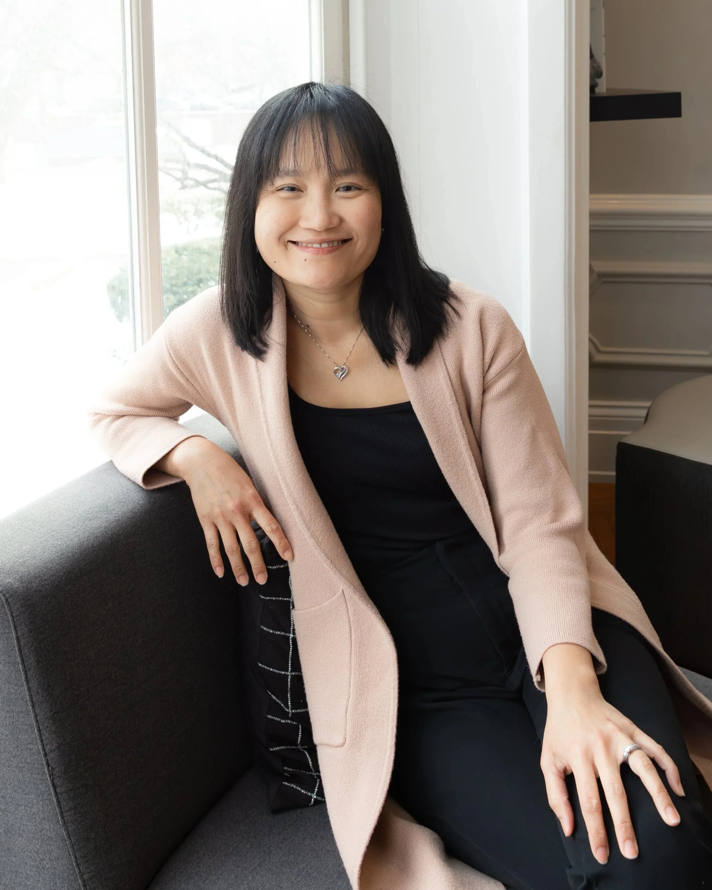 A woman sitting on a dark gray couch near a window with snow outside, wearing a light pink blazer over a black top, with black pants and a silver necklace with a heart pendant, smiling at the camera.