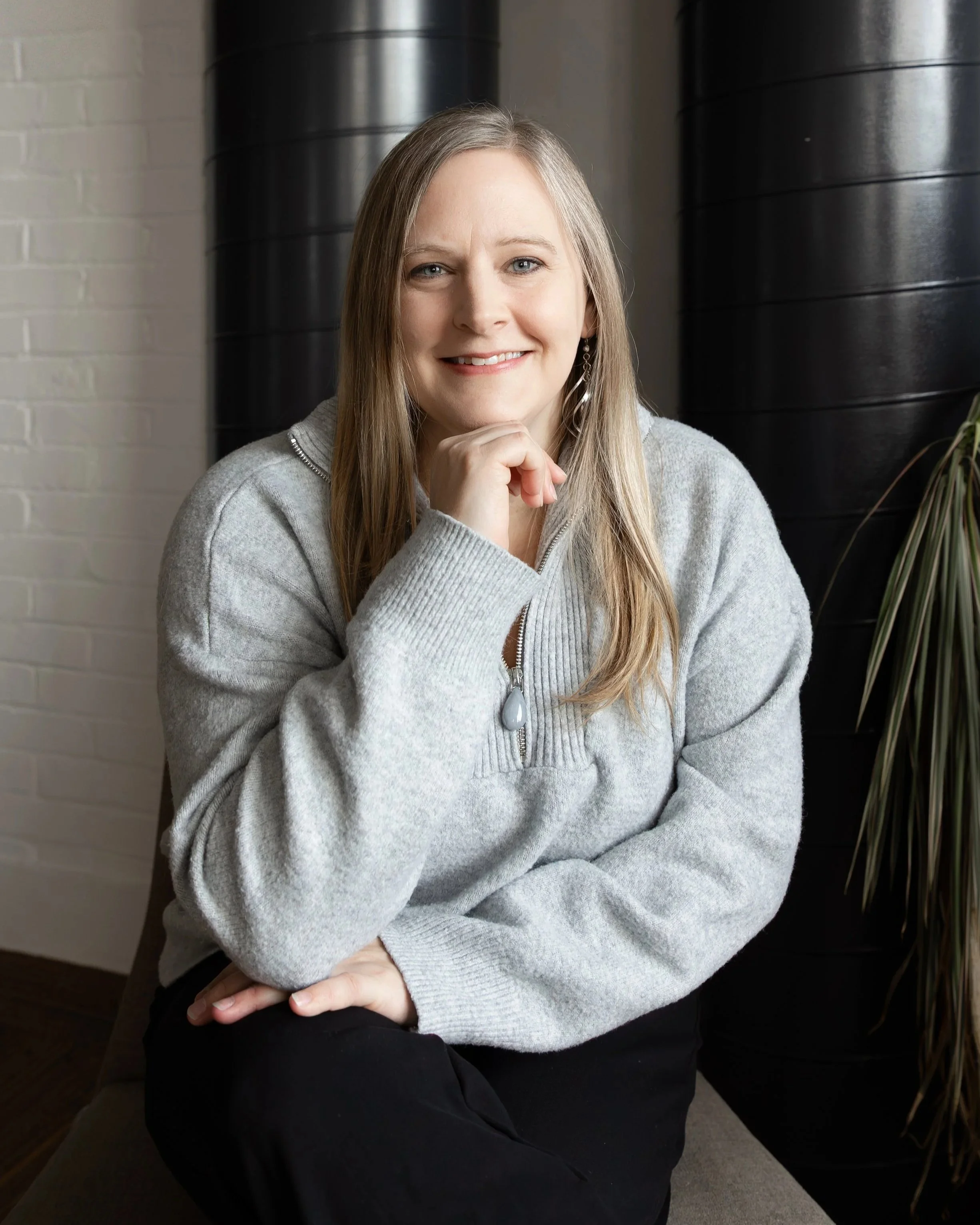 A woman smiling while holding an iPad, wearing a yellow blazer and light blue top, standing against a plain background.