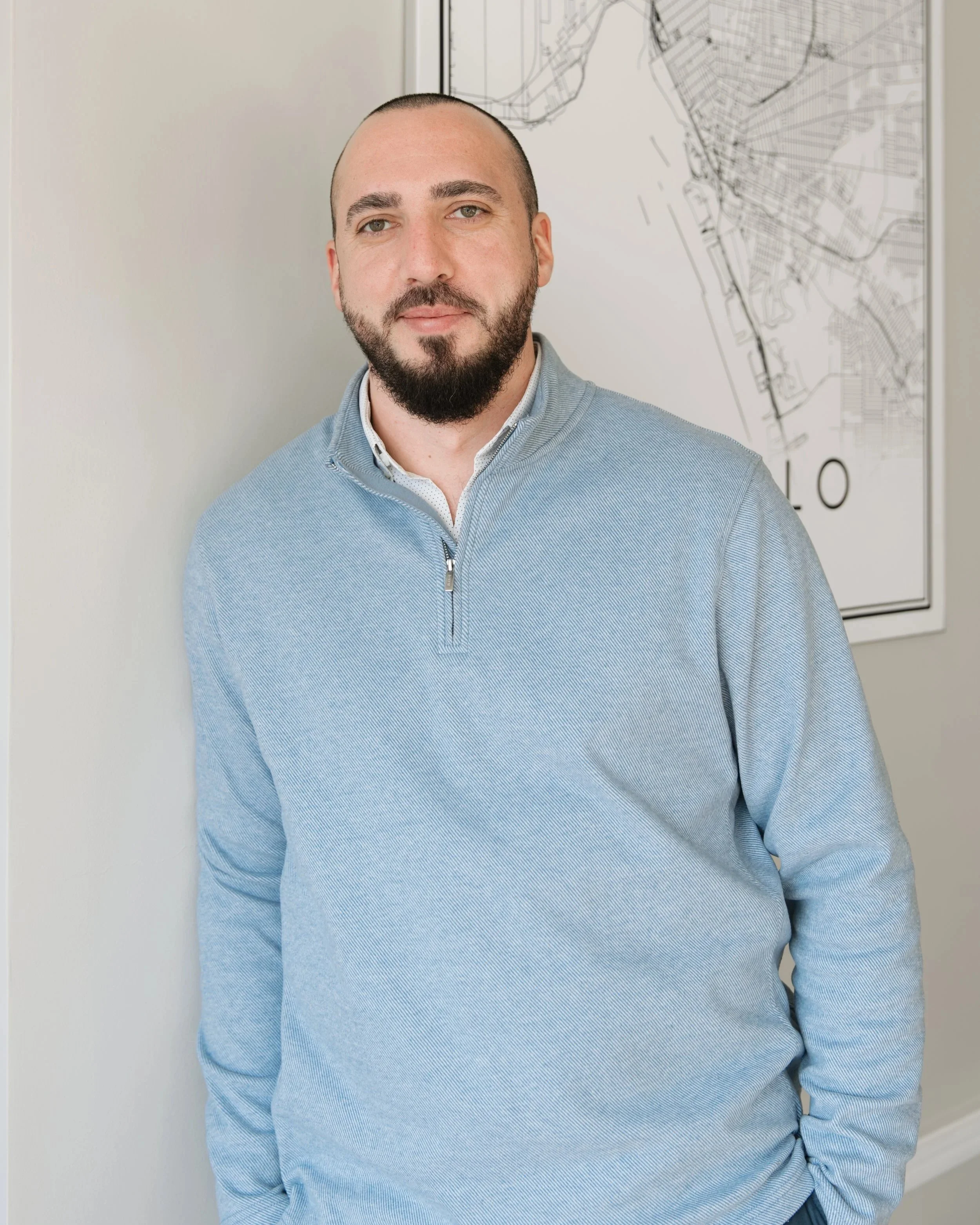 A man with a beard and short hair, wearing a light blue quarter-zip sweater over a collared shirt, stands indoors against a white wall with a large map behind him.