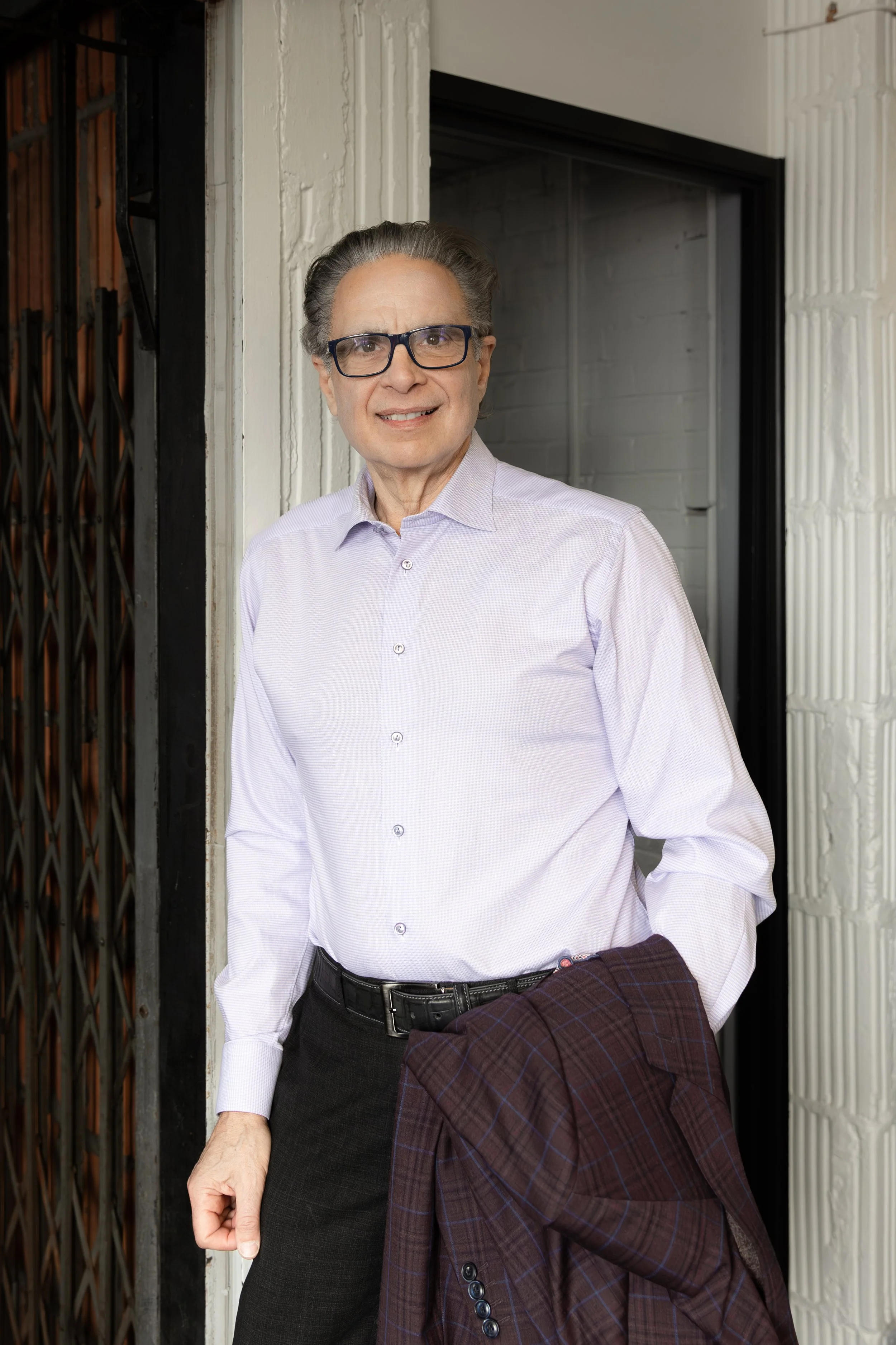 An older man with gray hair, glasses, wearing a light purple dress shirt, black trousers, and holding a plaid jacket. He is standing outside near a doorway and smiling at the camera.