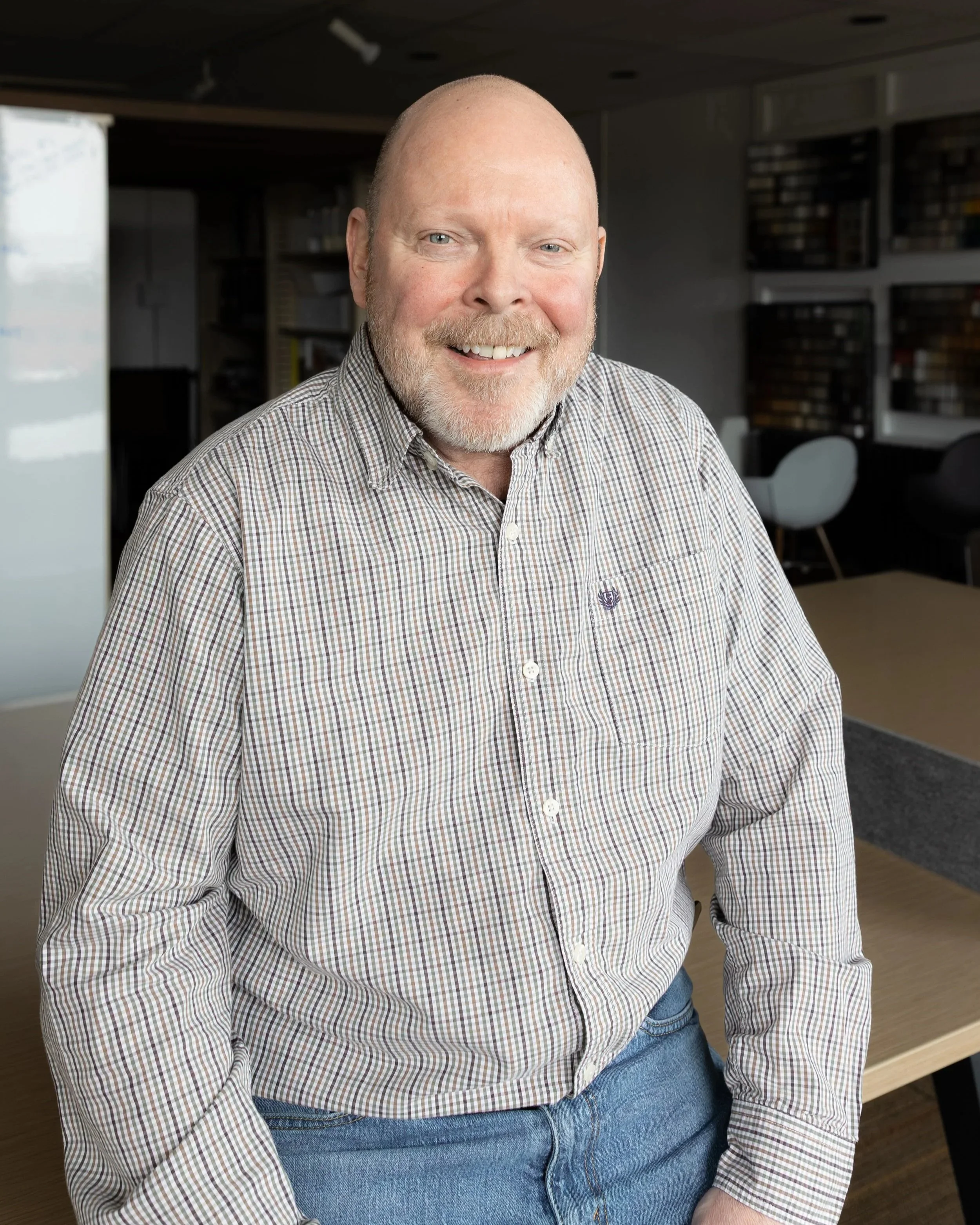 A smiling man with a beard and short hair standing against a light stone wall, wearing a black polo shirt and beige pants with a brown belt.