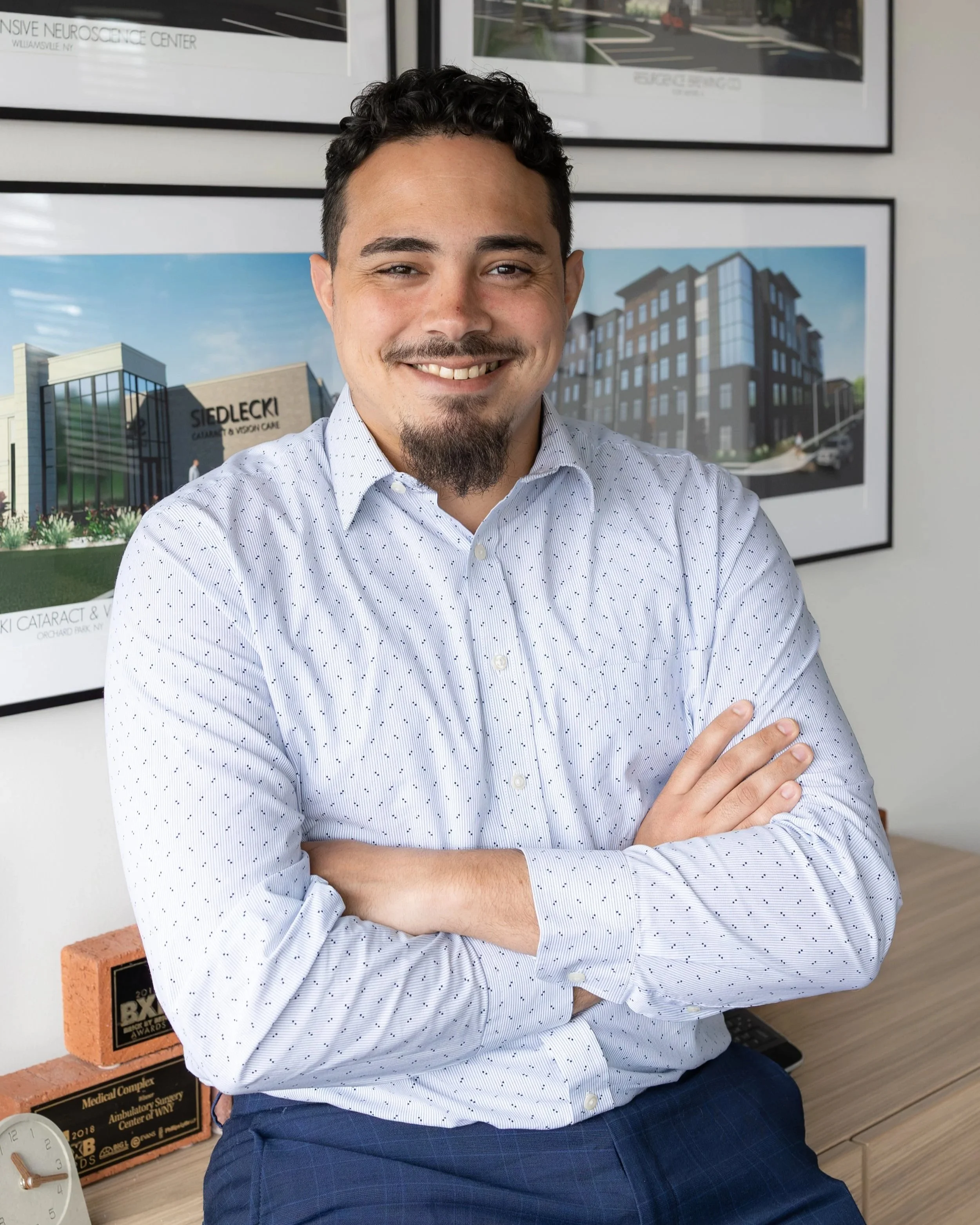 A smiling man with dark curly hair and a goatee, wearing a white polka dot dress shirt, standing with arms crossed in front of framed architectural drawings on the wall.