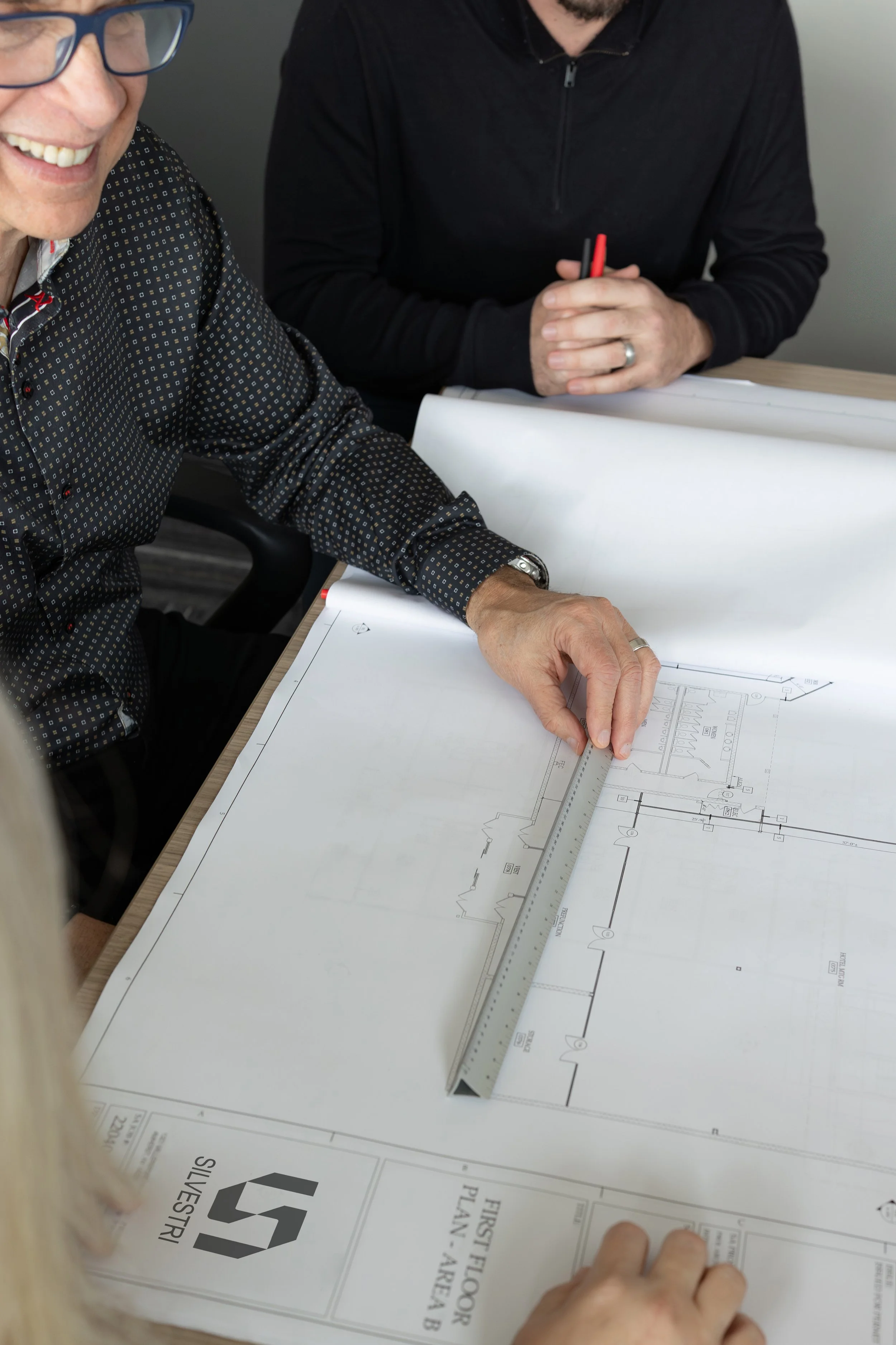 Two people reviewing a building blueprint, one using a ruler to measure the plan on a desk.