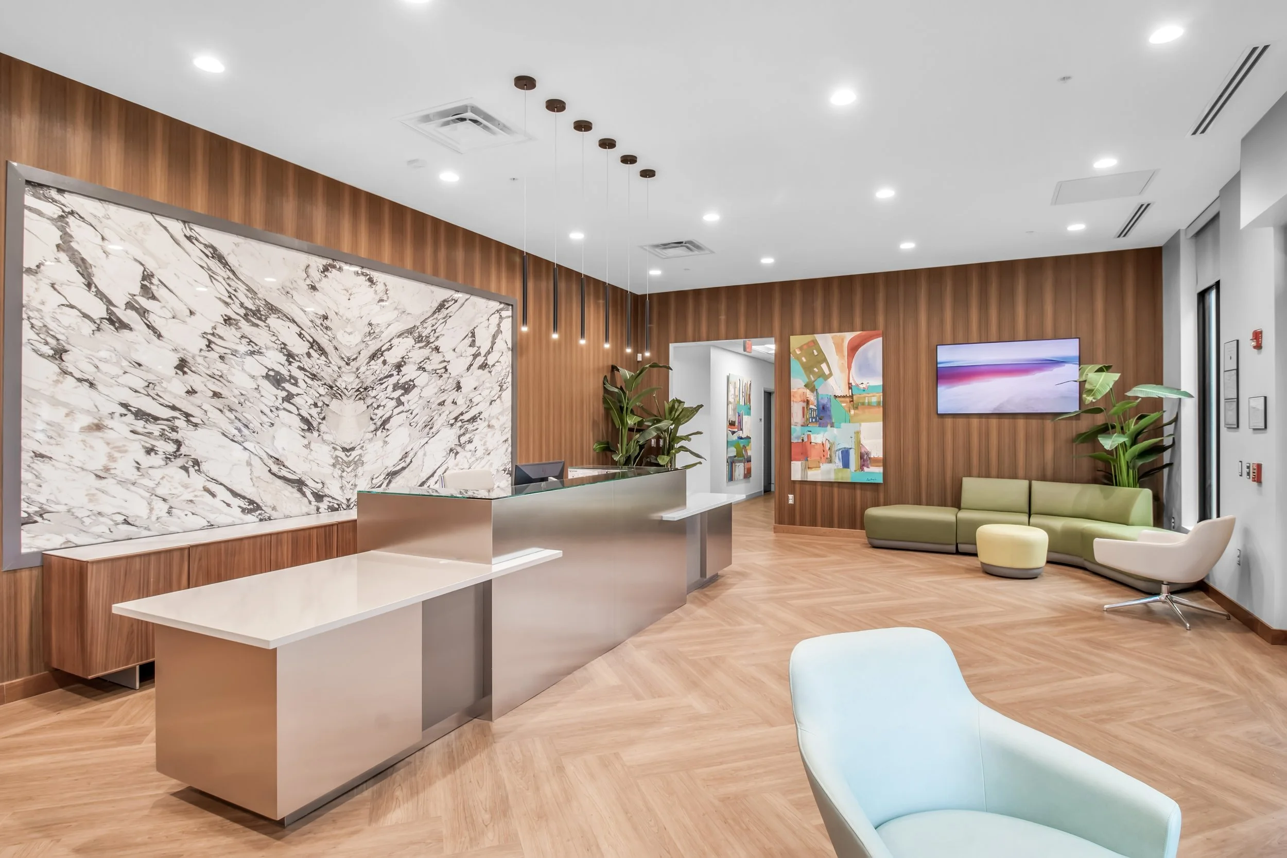 Modern lobby with wood-paneled walls, marble accent wall, reception desk, colorful artwork, green sofa, white lounge chair, TV, potted plants, and wood flooring.