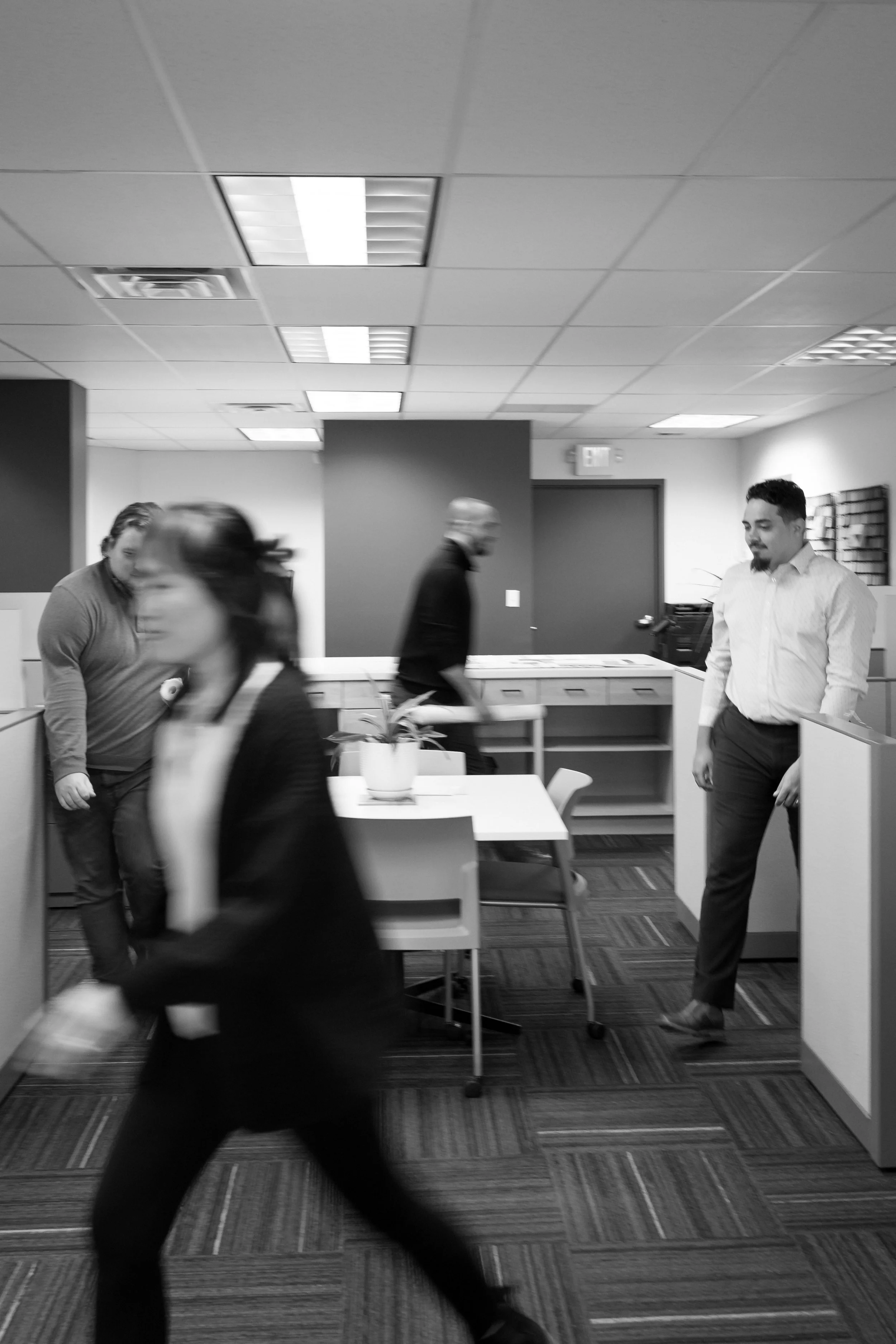 Black and white photo of an office space with four people. One woman in business attire is walking through the room, and three men are standing or walking, engaged in conversation or moving around. The office has ceiling lights, a table with a potted plant, and cubicles.