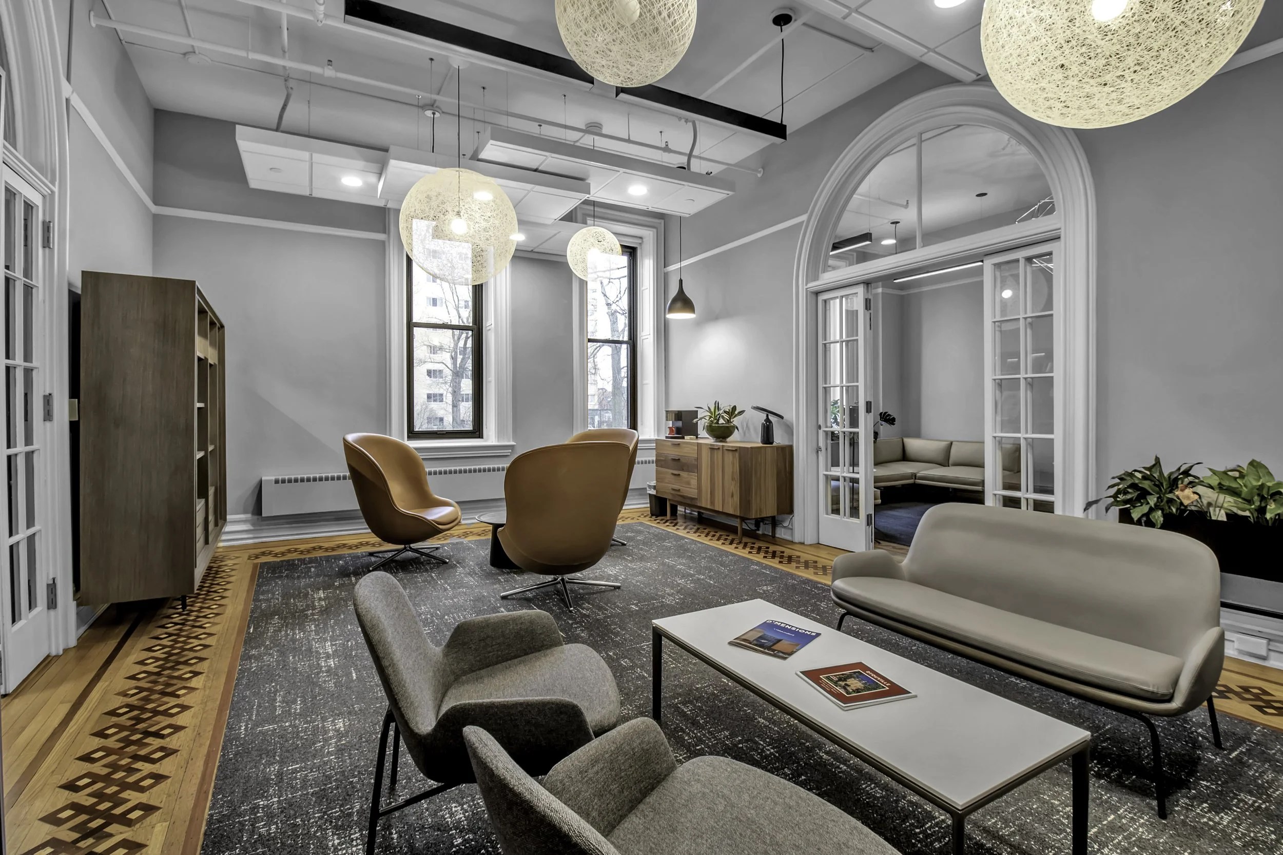 An interior lounge area with multiple chairs and sofas, wooden accent furniture, large windows, pendant lighting, plants, and a doorway leading to a separate seating room.