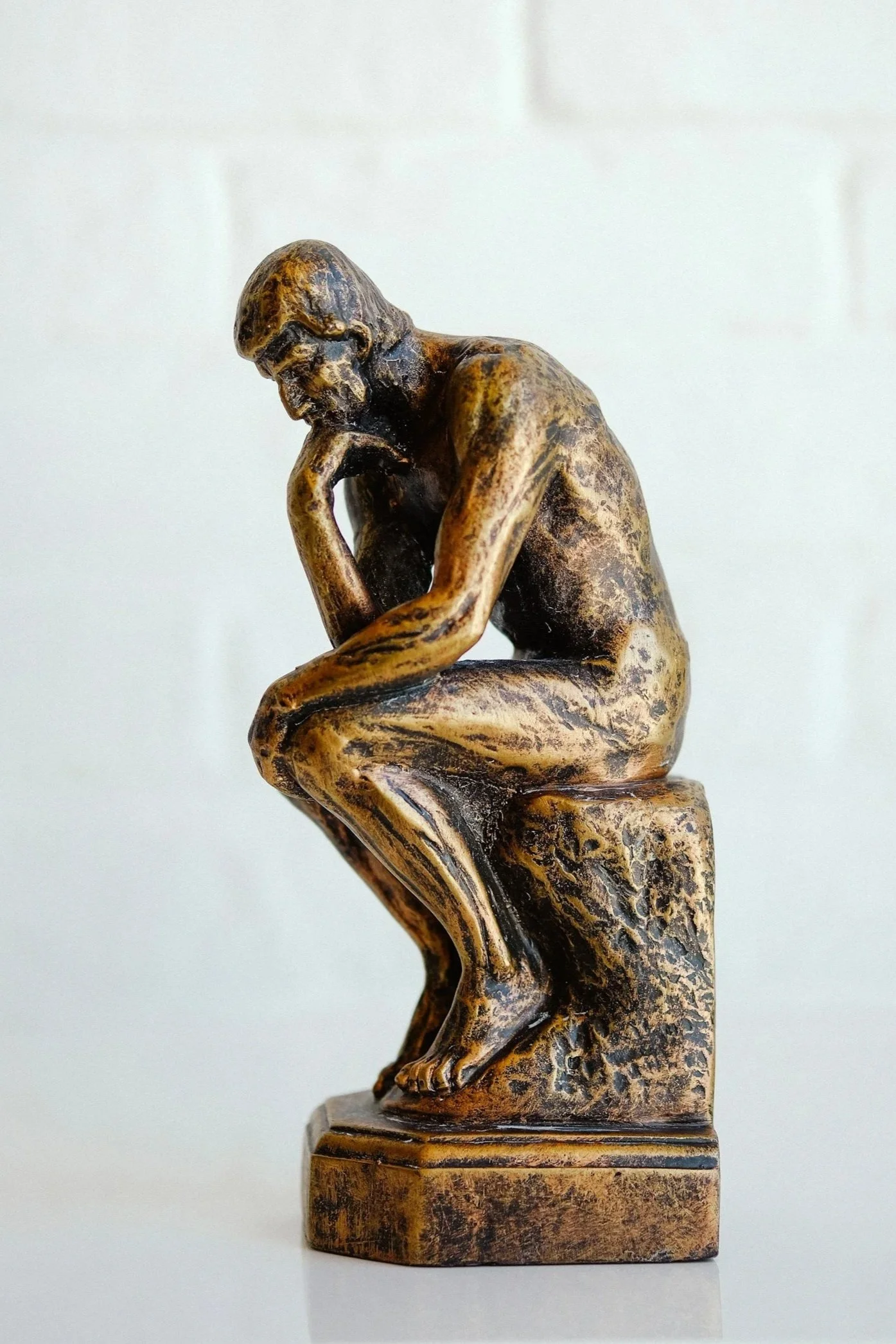 Bronze-colored sculpture of a man sitting on a block with his head resting on his hand, deep in thought, against a plain white background.