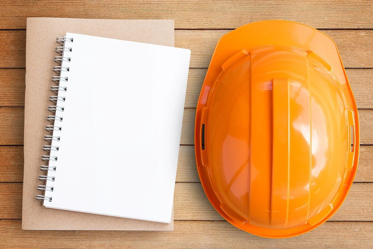 An orange construction helmet, a spiral-bound notebook, and a brown folder are placed on wooden planks.