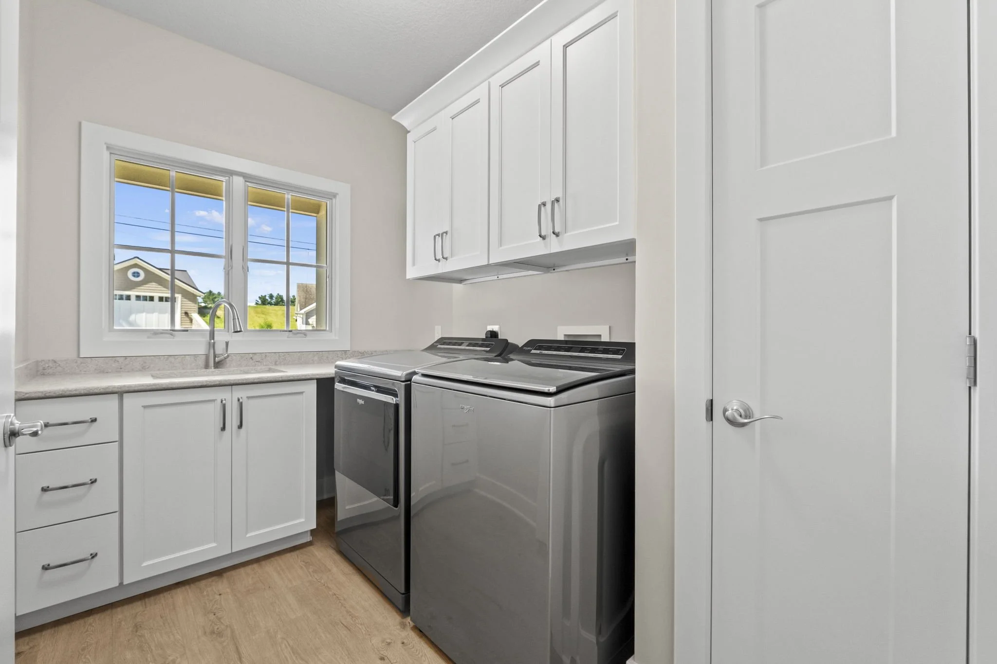 Laundry room with white cabinets, a window above the sink, a front-loading washing machine and dryer in silver, and wood flooring.