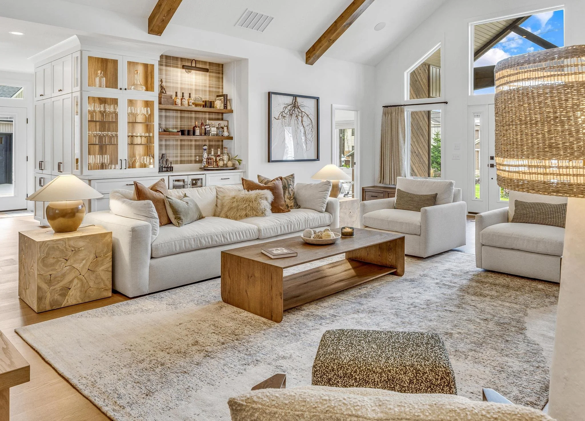 Bright living room with white sofa, armchairs, wooden coffee table, built-in bar, large windows, and decorative pillows.