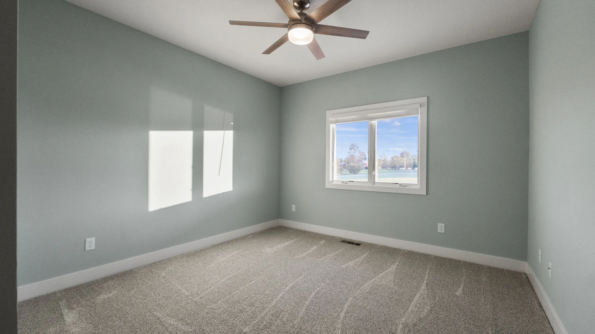 Empty bedroom with light green walls, beige carpet, a window showing trees and blue sky, and a ceiling fan with lights.