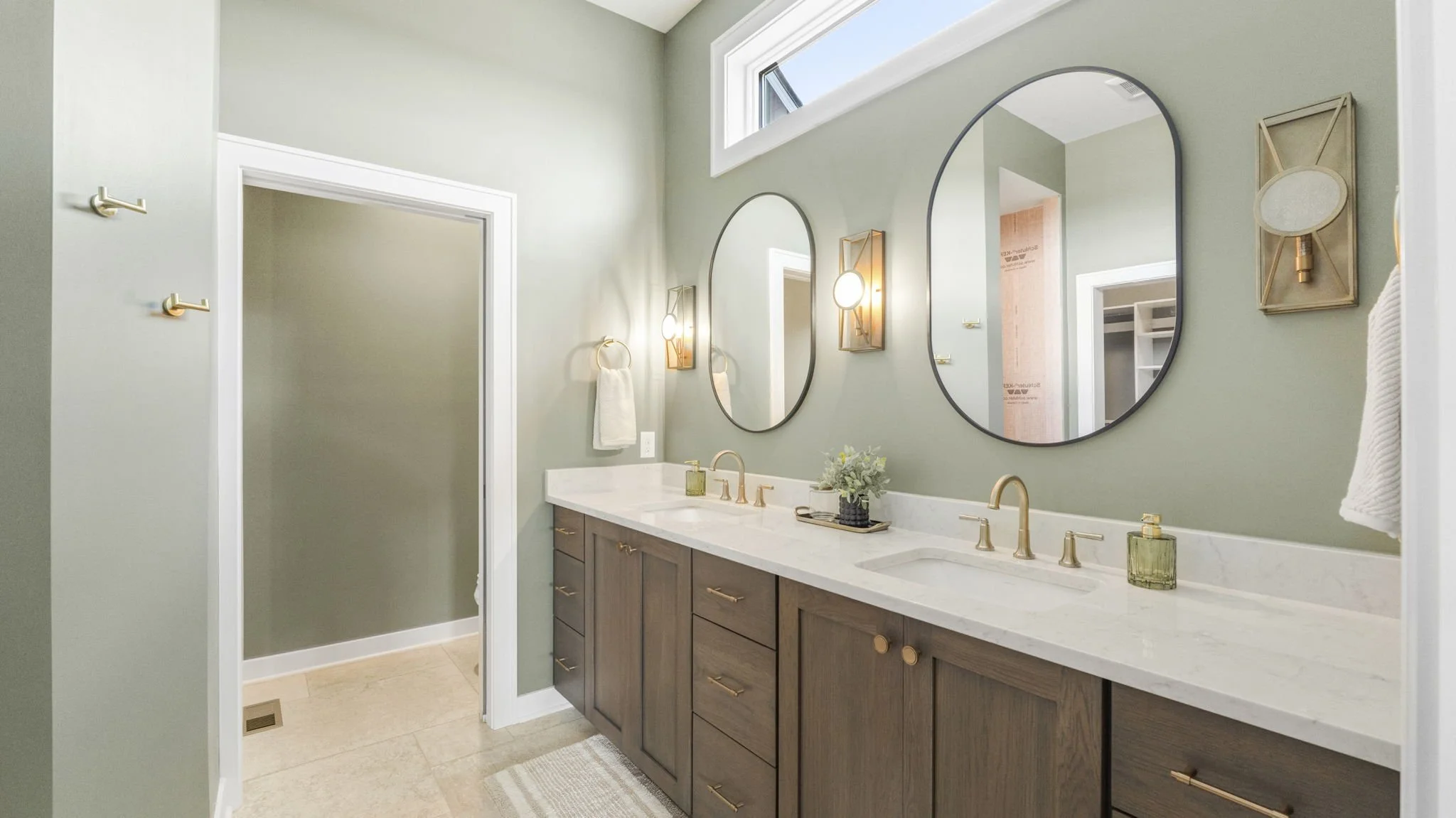 Modern bathroom with double vanity, two oval mirrors, gold fixtures, light green walls, and decorative wall sconces. There is a flower arrangement on the counter and a window above the mirrors.