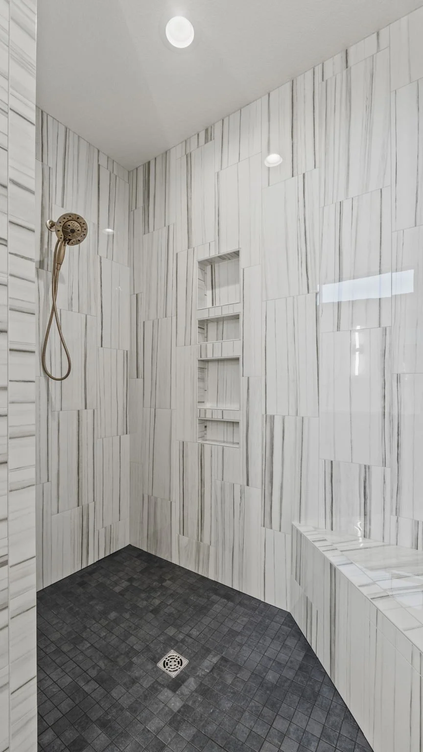 Empty modern shower with white marble walls, built-in shelves, a bronze showerhead, and black tile flooring.
