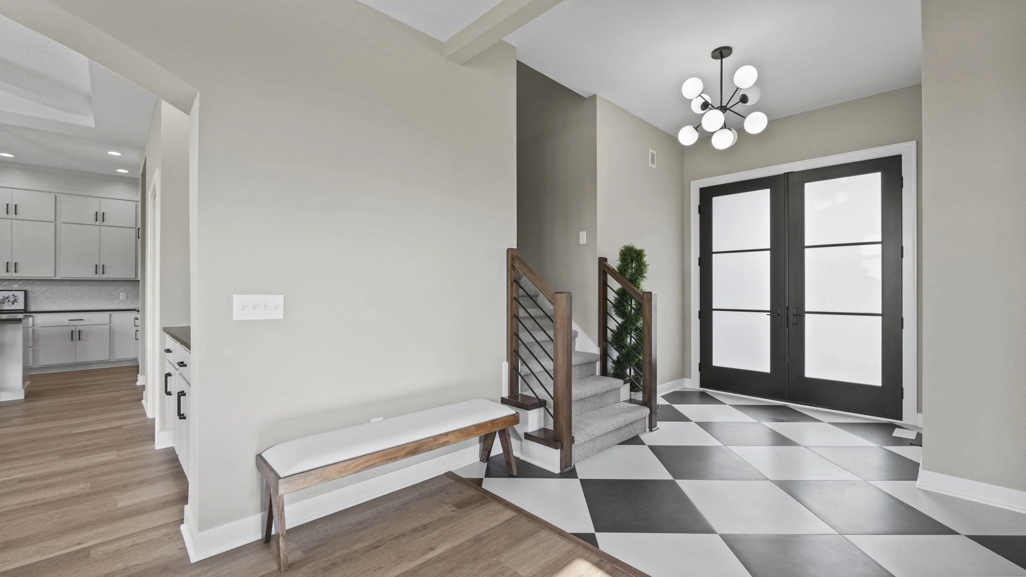 Modern entryway with black double doors with frosted glass, checkerboard-patterned tile flooring, a small staircase with wooden railing, a sitting bench, and a contemporary chandelier.