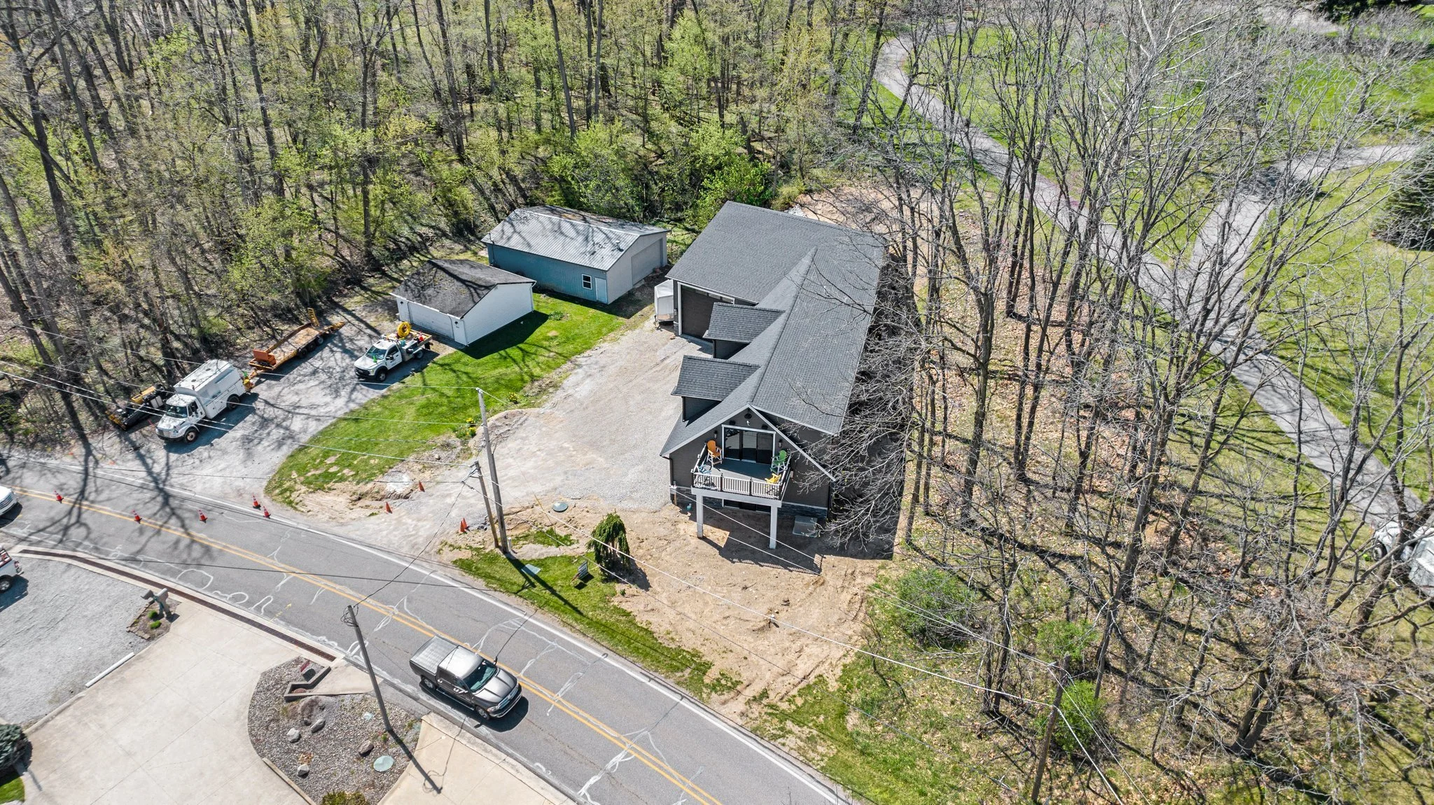 Aerial view of a residential house with a deck, surrounded by trees, with parked cars and construction equipment nearby.