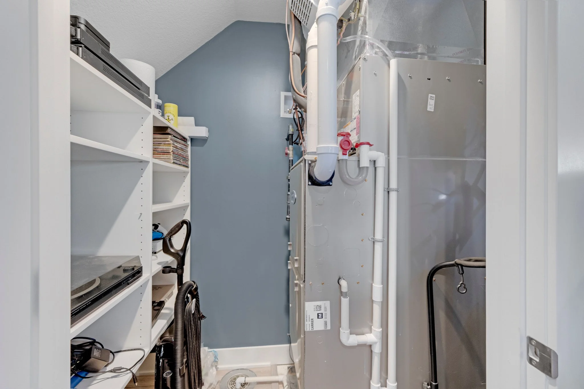 A utility closet with a white shelving unit on the left holding vinyl records, a turntable, and various electrical items; the center features HVAC equipment with white pipes and wiring; the right side shows a shiny metal surface and a black walker.