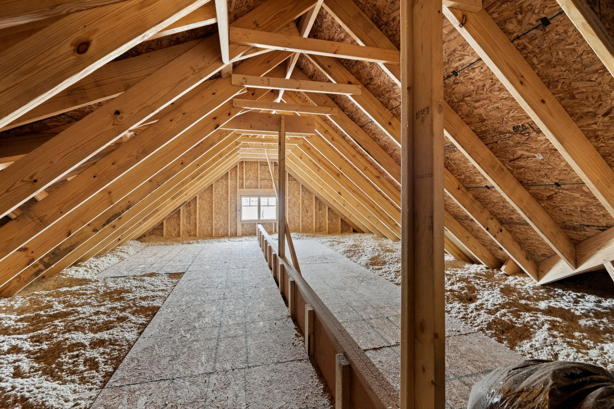 Attic with wooden roof trusses, insulation on the floor, and a small window at the far end.