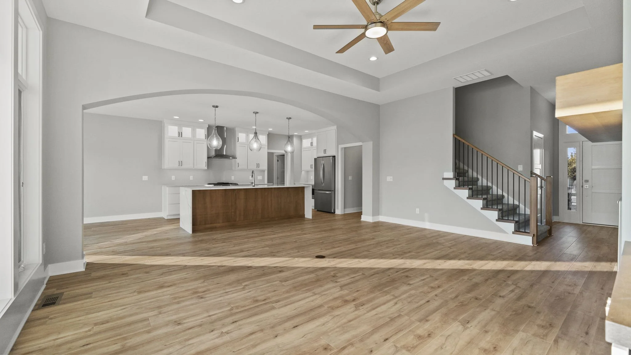 Open-concept living space with hardwood floors, a kitchen with white cabinets, a large island with wood paneling, stainless steel appliances, three pendant lights, a staircase with a wooden railing, and a ceiling fan with wooden blades.