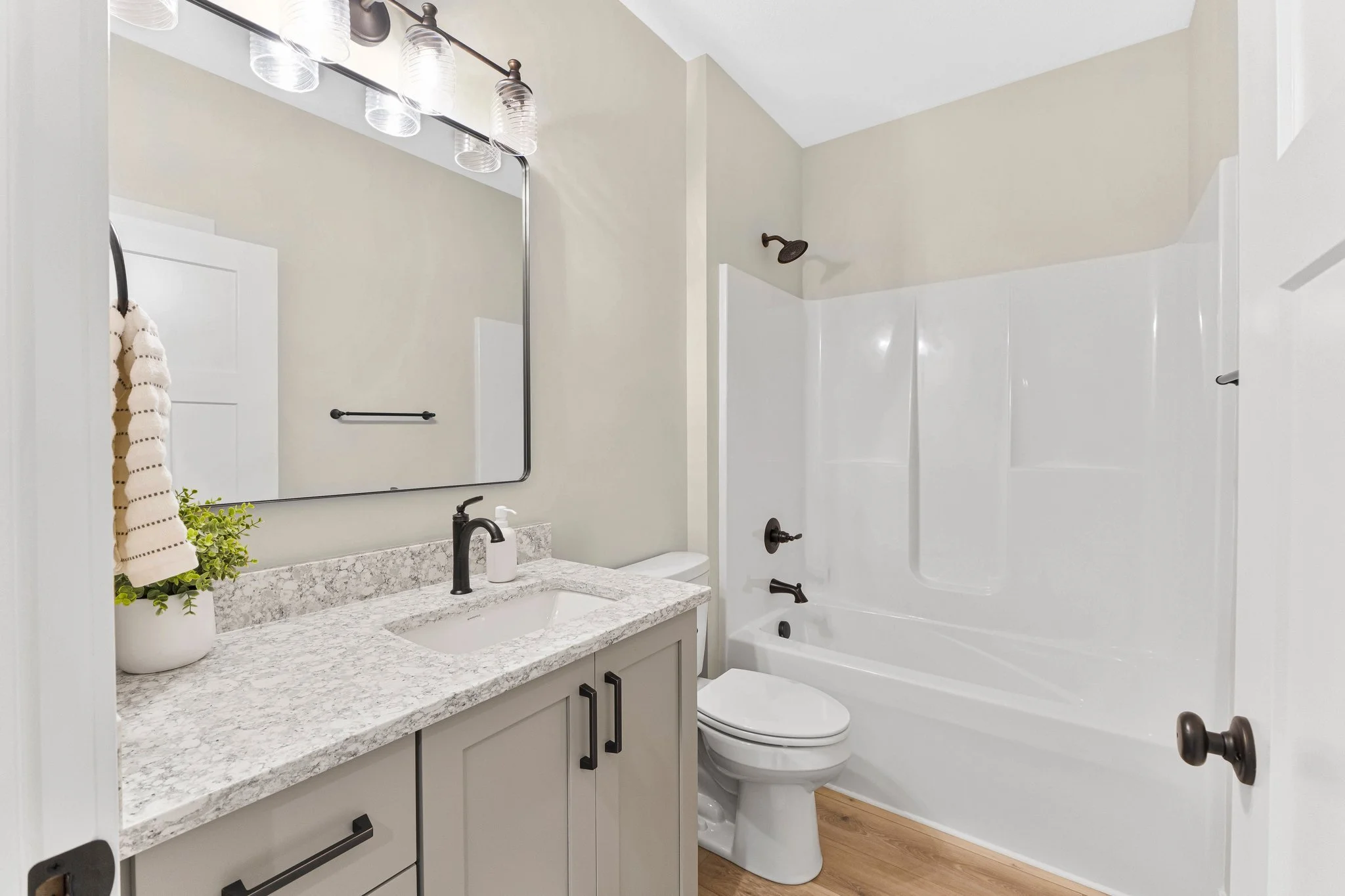 Clean bathroom with beige walls, white bathtub with shower, toilet, granite countertop vanity with a plant and soap dispenser, large mirror, and wooden flooring.