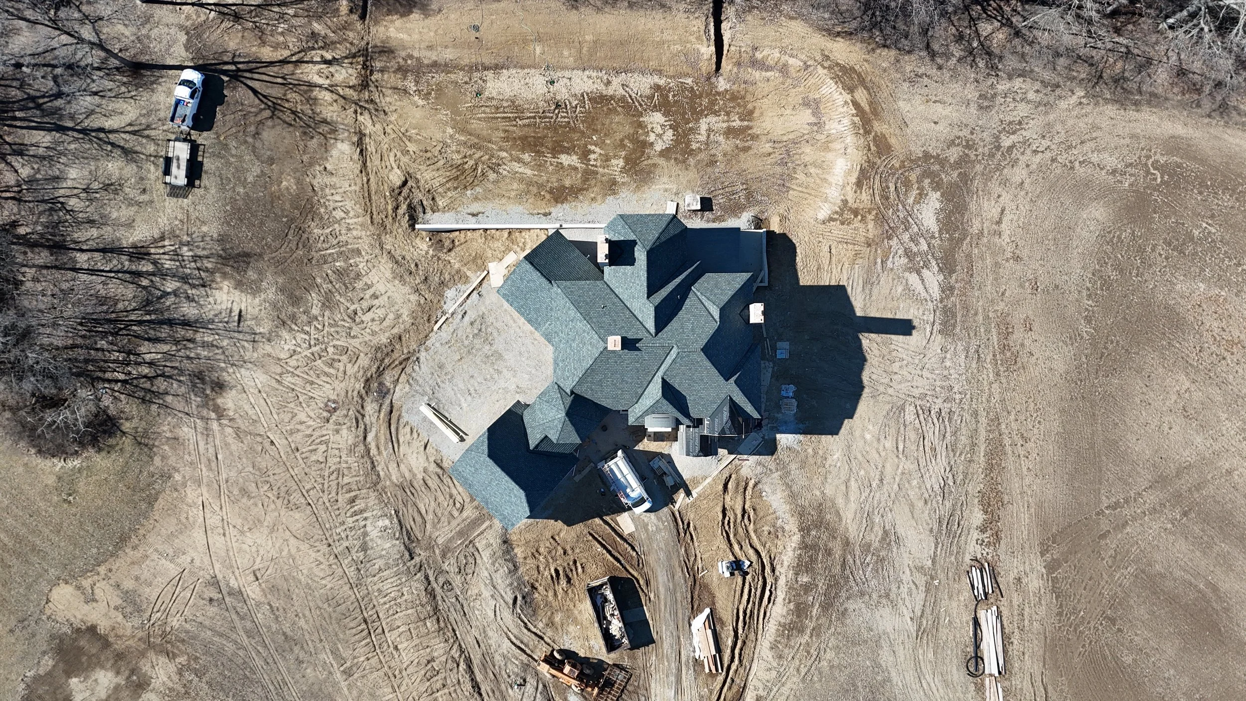 An aerial view of a house under construction in a large, excavated lot with construction vehicles and materials nearby.