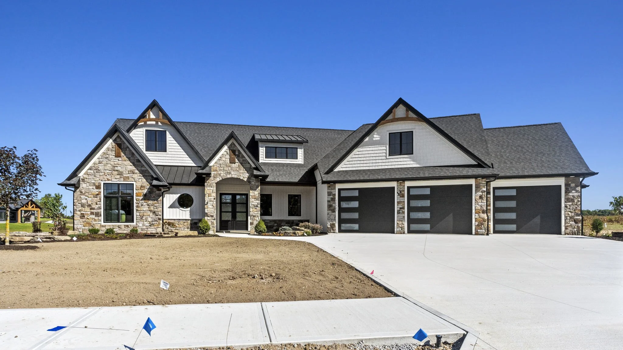 Newly constructed modern house with stone and white siding exterior, black garage doors, and a large concrete driveway under a clear blue sky.