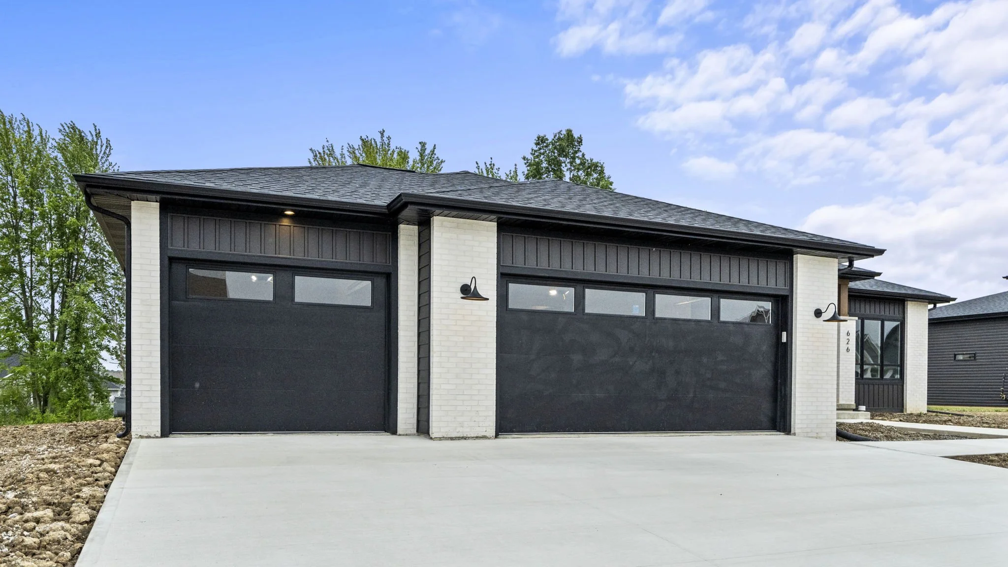 Modern house with white brick walls, black garage doors, and black accent trim, with a concrete driveway and green trees under a partly cloudy sky.