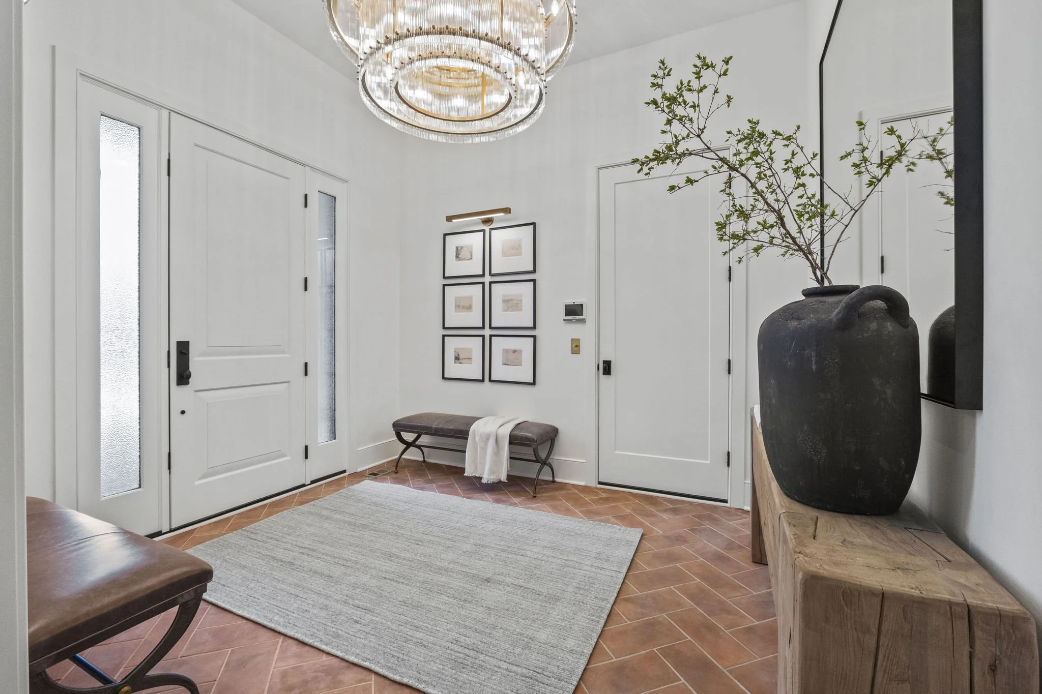 Modern entryway with white walls, a large black vase with greenery on a wooden console, a gray rug, a framed photo gallery on the wall, and a bench with a throw blanket. The space features a statement chandelier and terracotta tile flooring.
