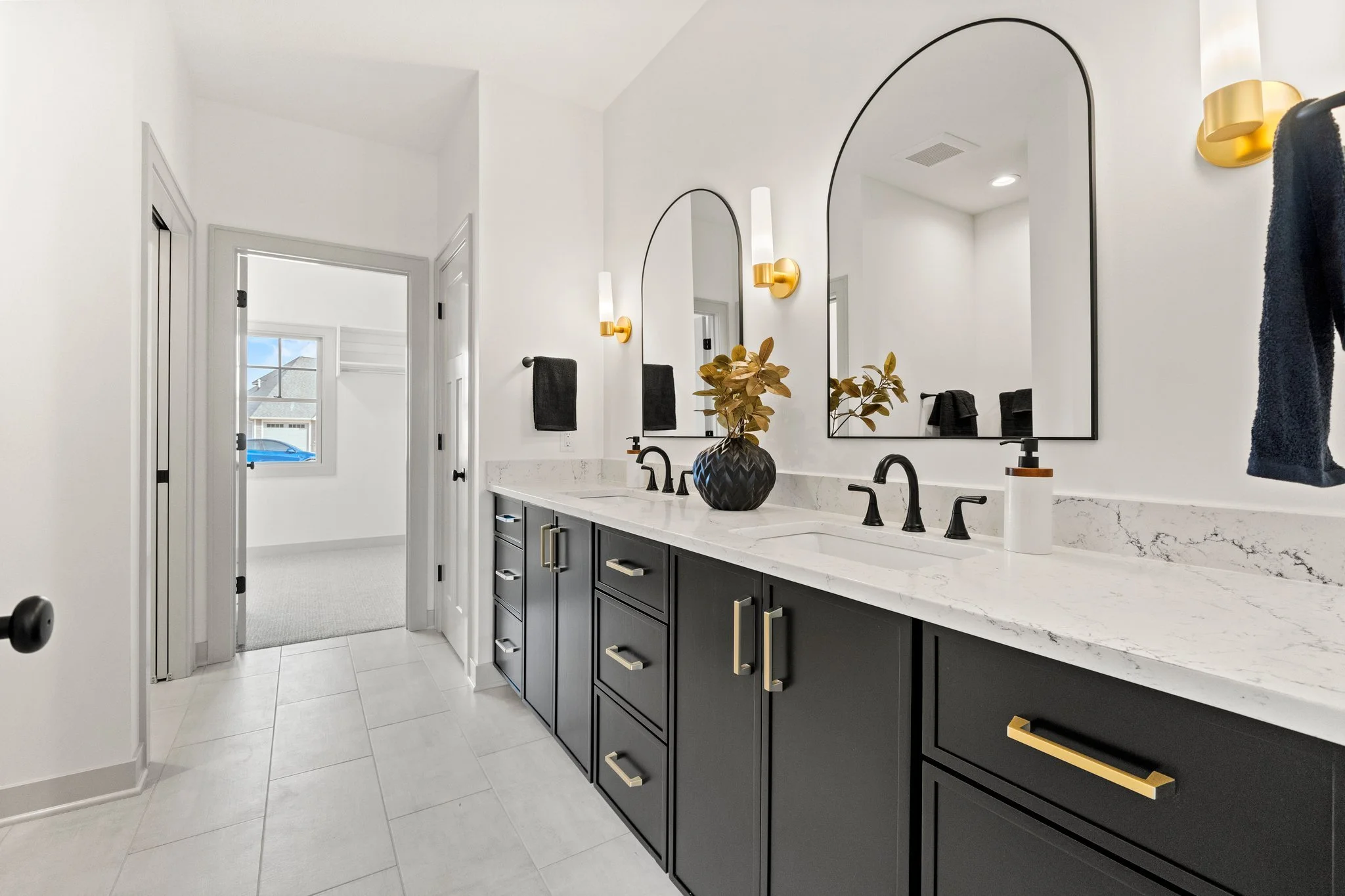 Modern bathroom with dark cabinets, white marble countertop, double sinks, oval mirrors, black fixtures, and gold accents. A black vase with gold leaves is on the counter, and there are gold wall sconces and black towels.