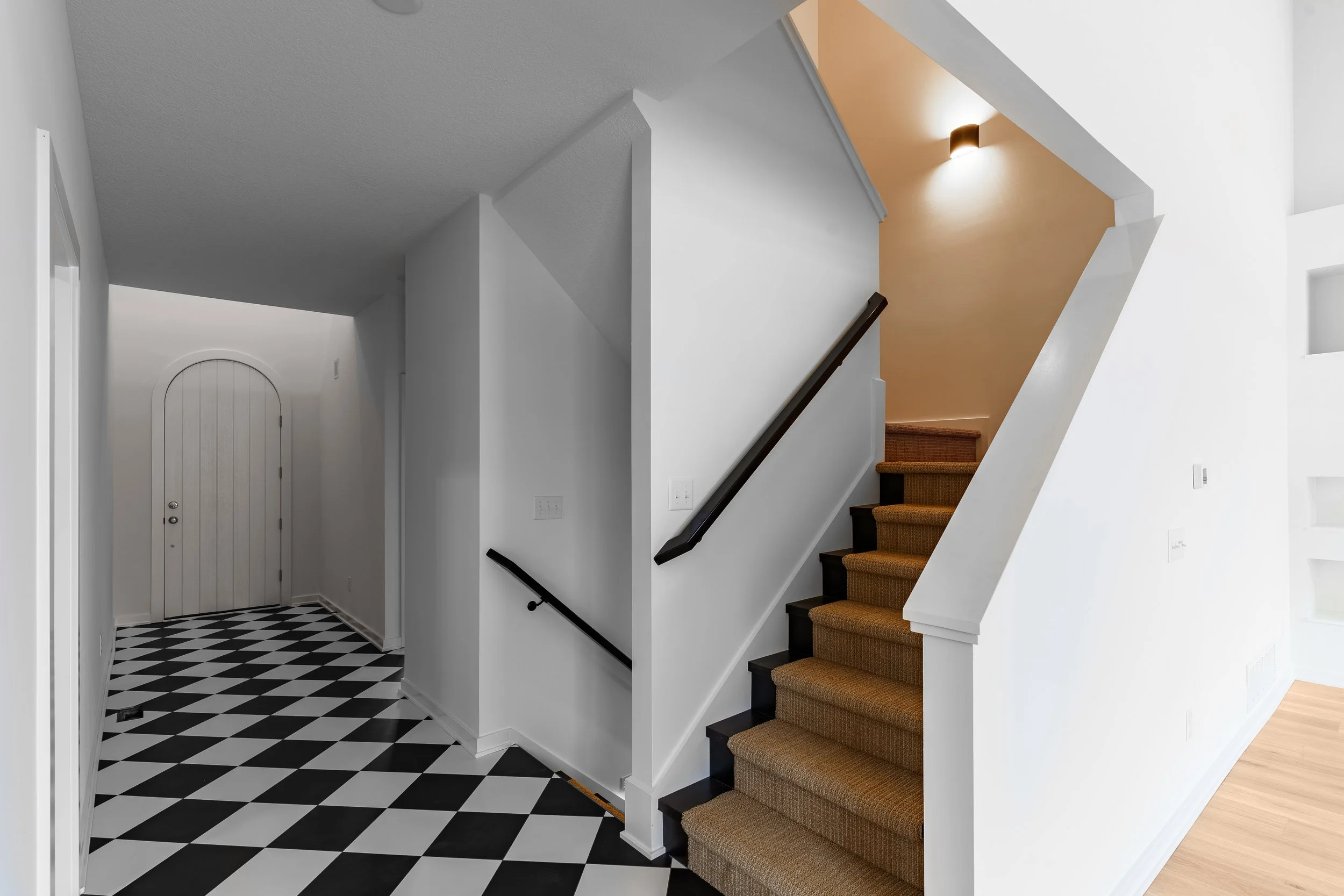 Interior of a house showing a staircase with brown carpet leading upstairs, a black handrail, white walls, and a hallway with a black and white checkered floor near a white arched door.