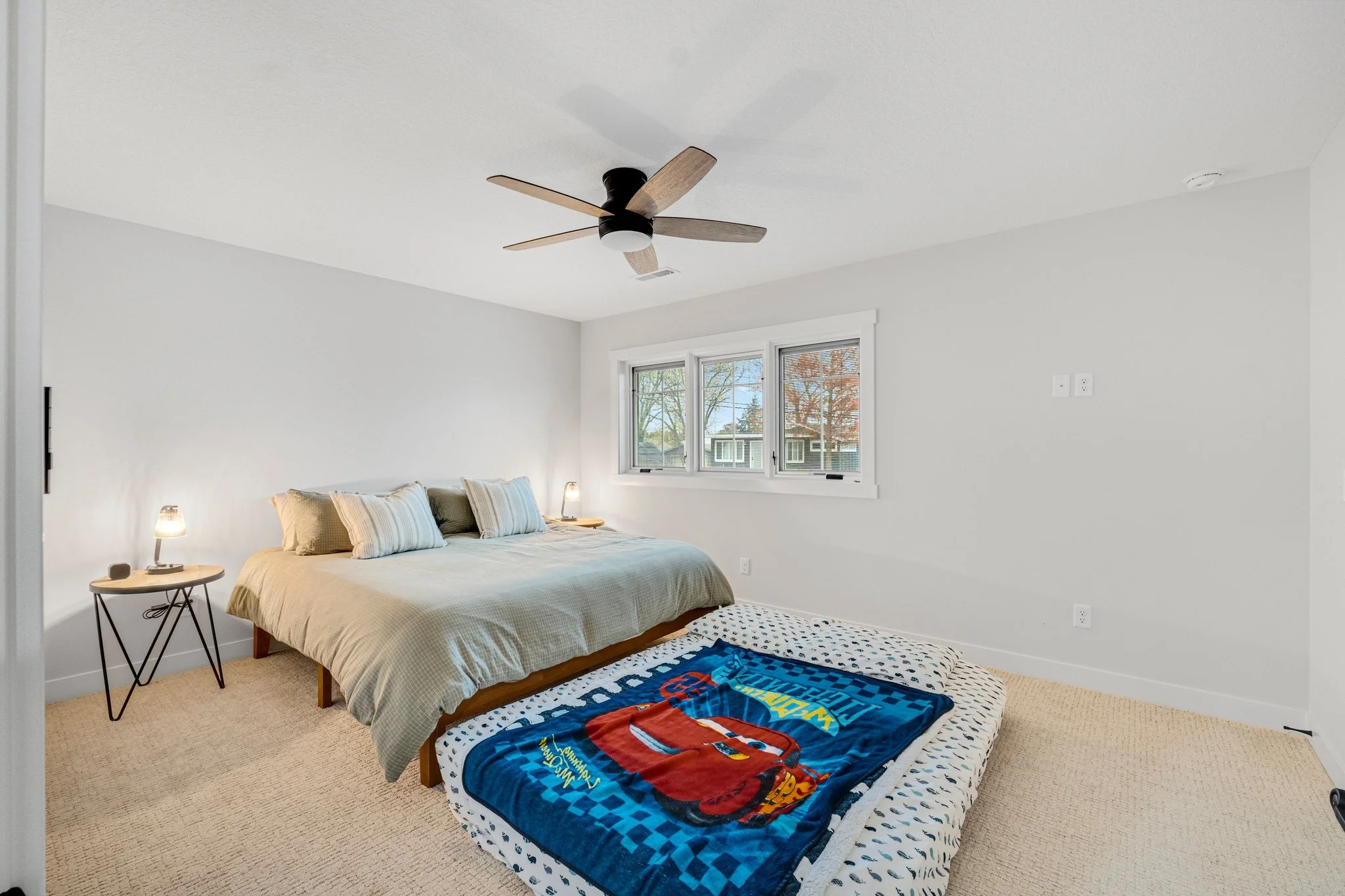 A bedroom with a bed, nightstands, and lamps, featuring a Car's Lightning McQueen blanket on a mattress on the floor, with a window showing trees outside.